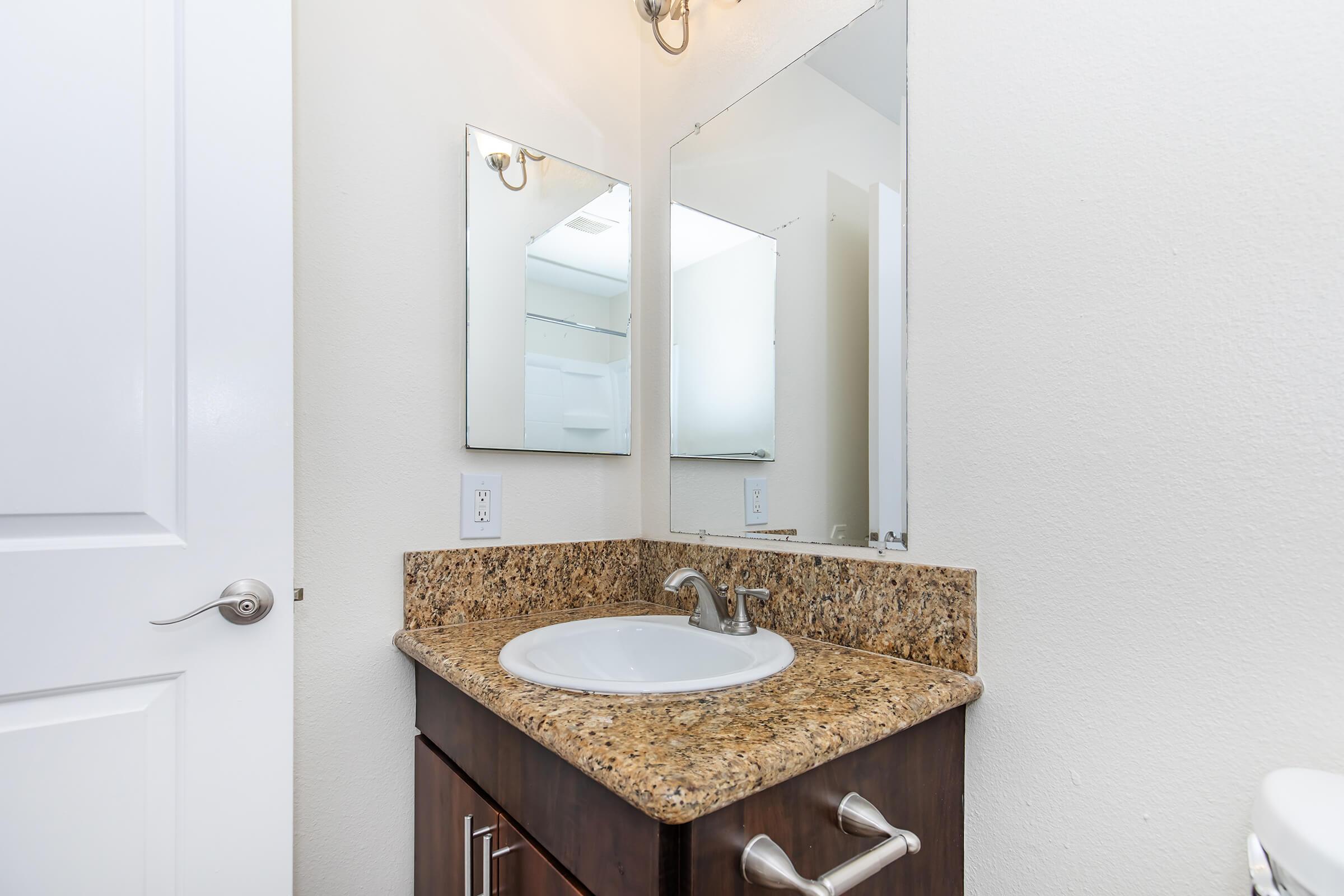 A modern bathroom with a granite countertop, a sink, and two mirrors mounted on the wall. The cabinetry is dark wood, and there is a light fixture above the mirrors. The walls are painted white, and a white toilet is partially visible on the right. Overall, the space appears clean and well-maintained.