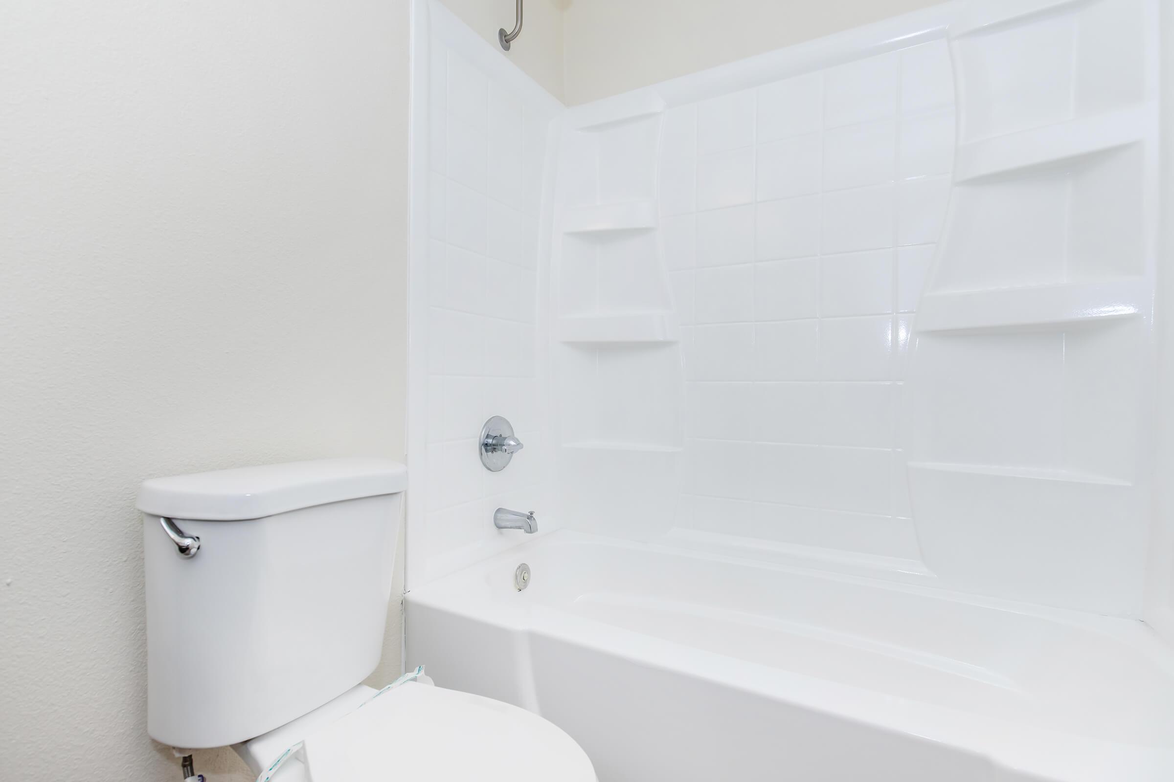 A clean, modern bathroom featuring a white bathtub with a shower, a toilet, and minimalist wall design in a light color. The tub and toilet are both installed against a plain wall, showcasing a simple and functional layout.