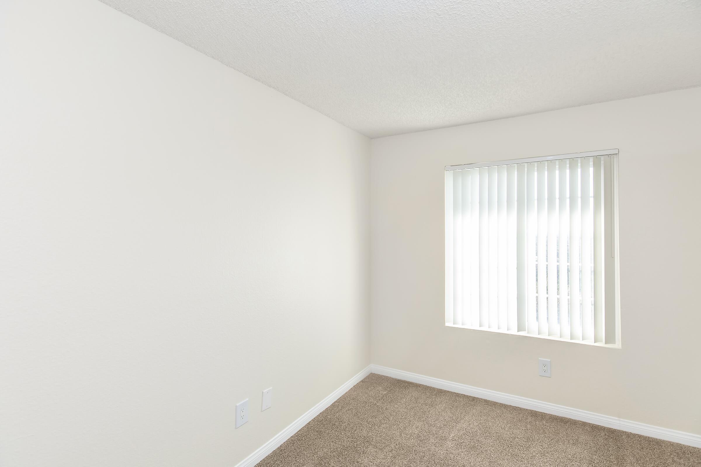 A small, empty room with light beige walls and a window covered with vertical blinds. The floor is carpeted in a light, neutral color, and there are two electrical outlets visible on one wall. The room appears bright and uncluttered, ideal for various uses.