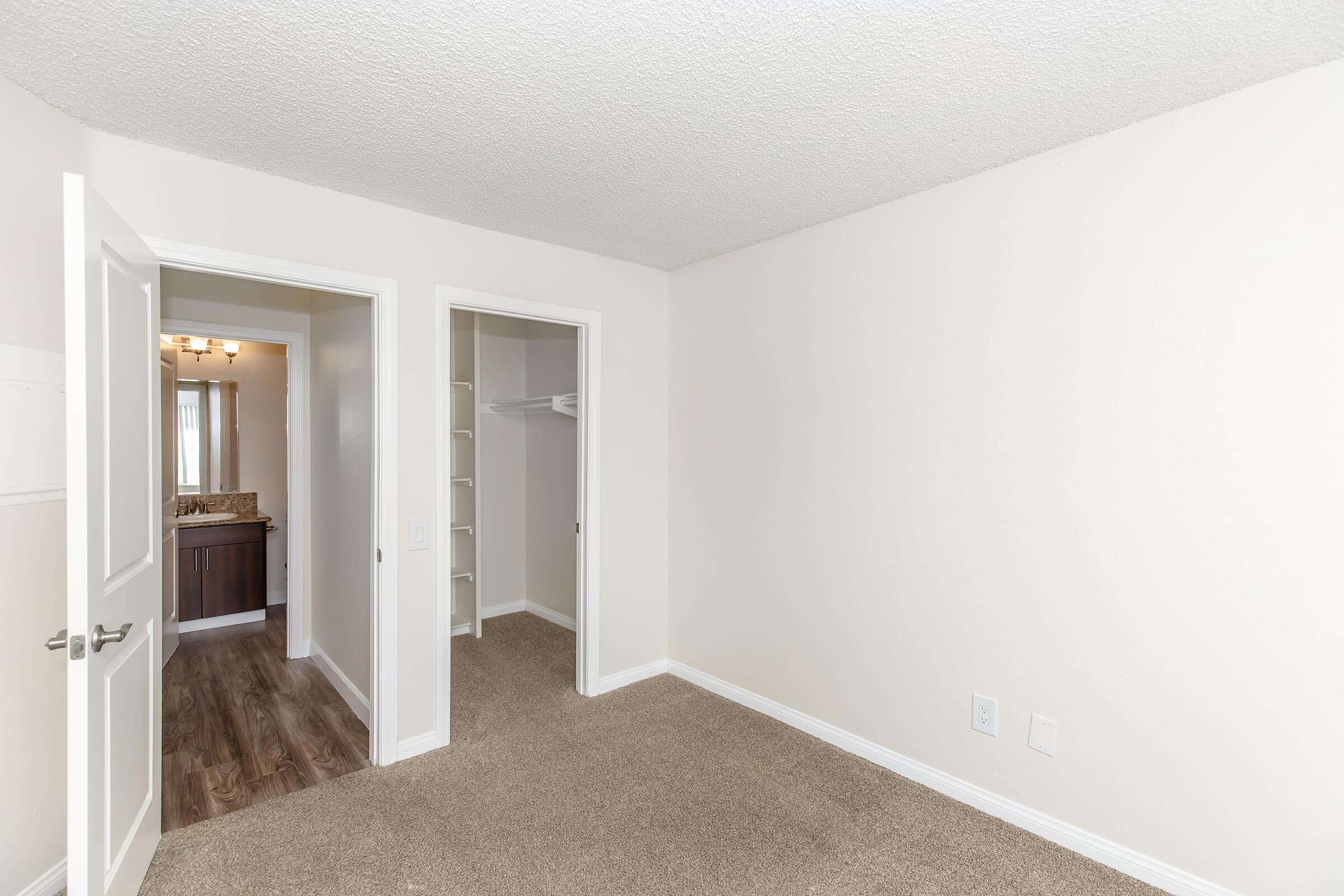 A well-lit, empty bedroom featuring beige walls and carpet. The room has an open door leading to a bathroom area, and two closets—one with a hanging rod and the other with built-in shelves. The overall atmosphere is clean and minimalistic, ideal for personalization.