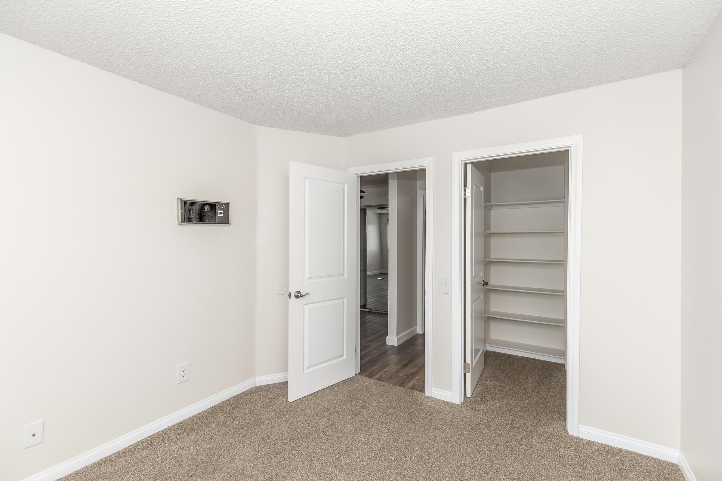 A well-lit, empty room featuring beige carpet and neutral-colored walls. There are two doors: one leading to a closet with shelves and the other to an adjacent room. A small wall-mounted clock or thermostat is visible on the left wall. The overall atmosphere is clean and spacious.