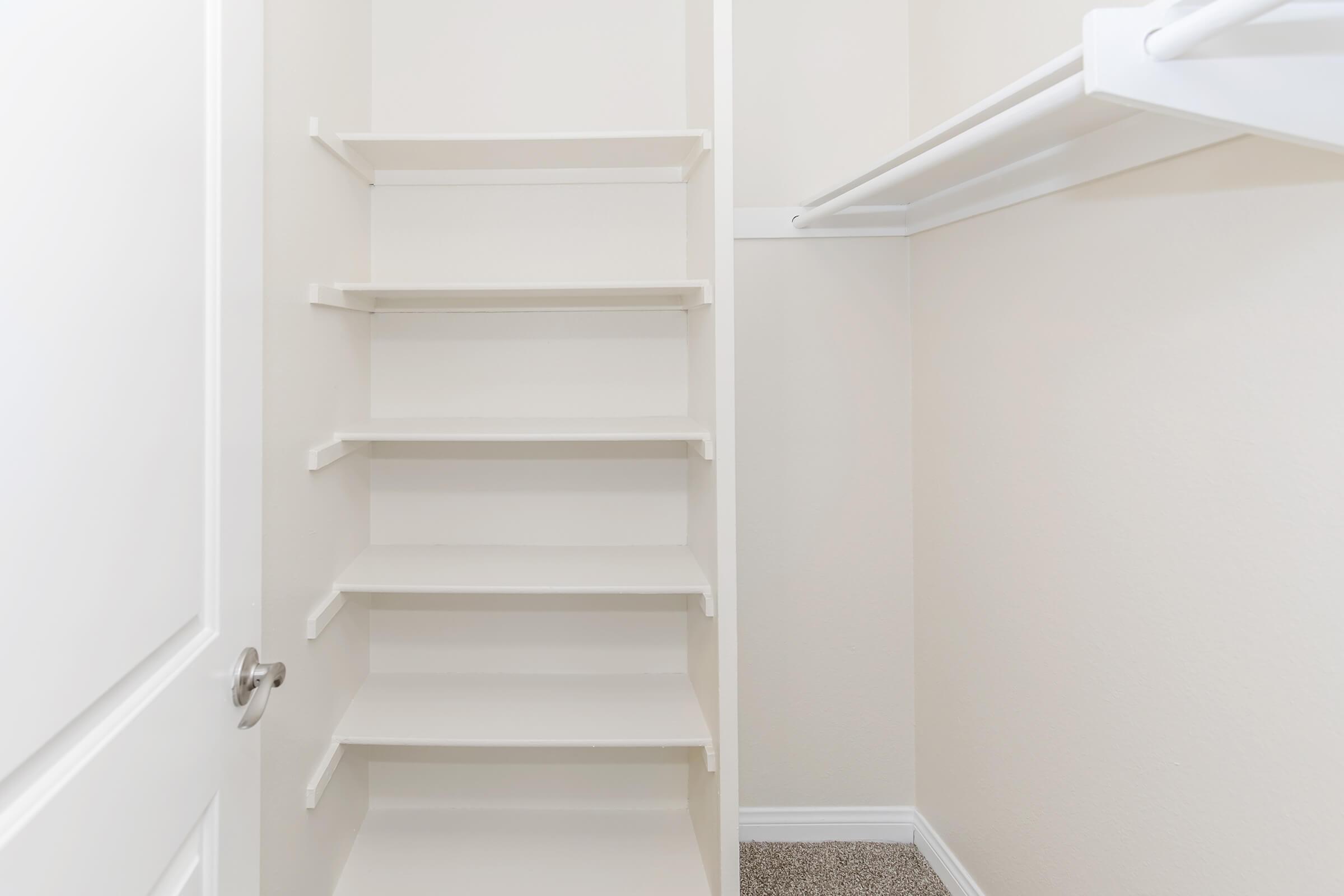A clean, empty closet featuring light-colored walls and a set of white shelves positioned on one side. There is also a hanging rod for clothes. The closet has a door with a simple handle, and the floor is carpeted in a soft beige color.
