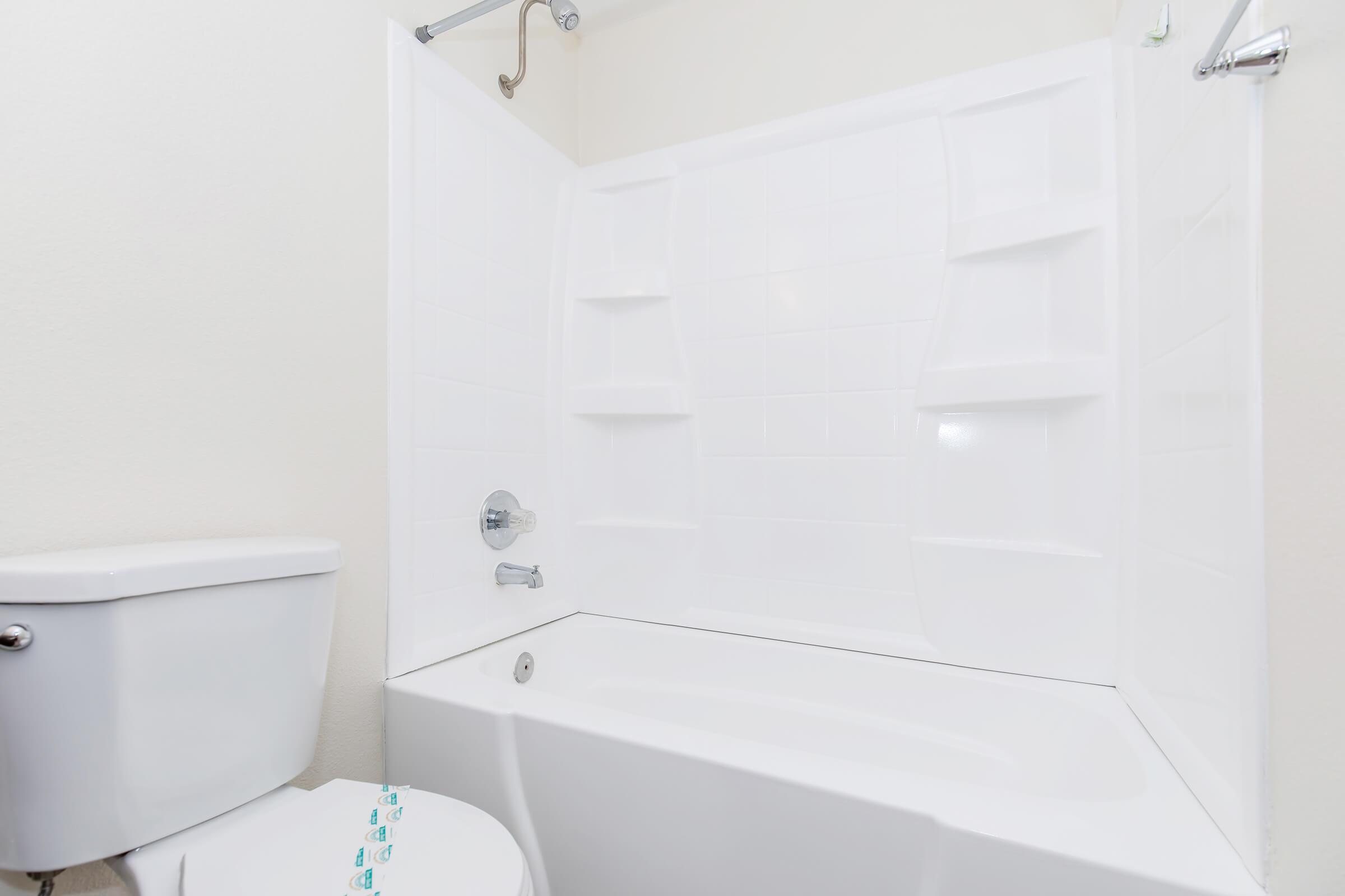 A clean, modern bathroom featuring a white bathtub with a showerhead, surrounded by white tiled walls. A white toilet is positioned next to the bathtub, with minimalistic decor and neutral colors. The overall look is simple and uncluttered, creating a bright and spacious atmosphere.