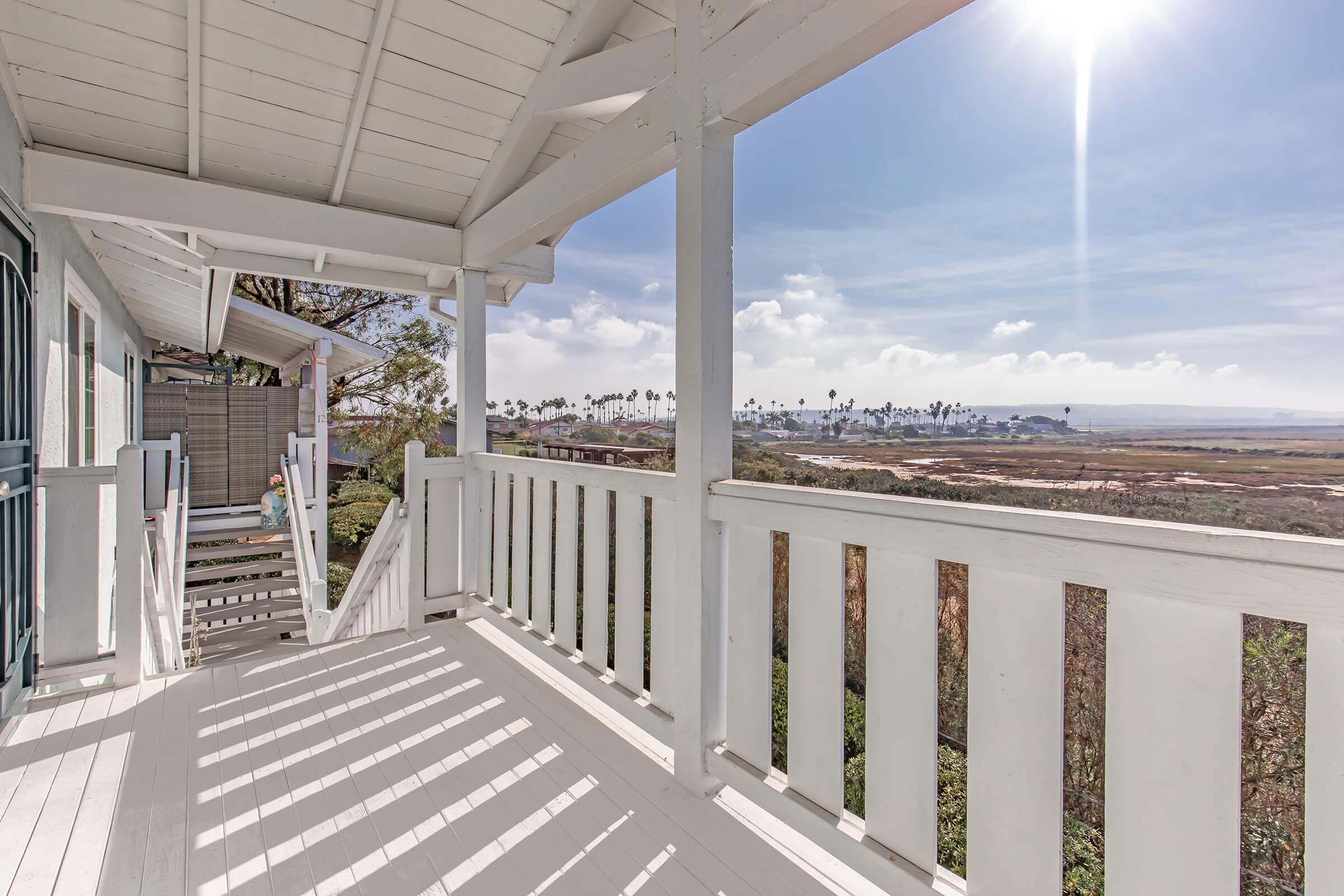 A view from a balcony overlooking a scenic landscape with palm trees and a clear sky. Sunlight filters through the railing, casting shadows on the white floor. The area below features a mixture of greenery and open space, creating a tranquil, inviting atmosphere.