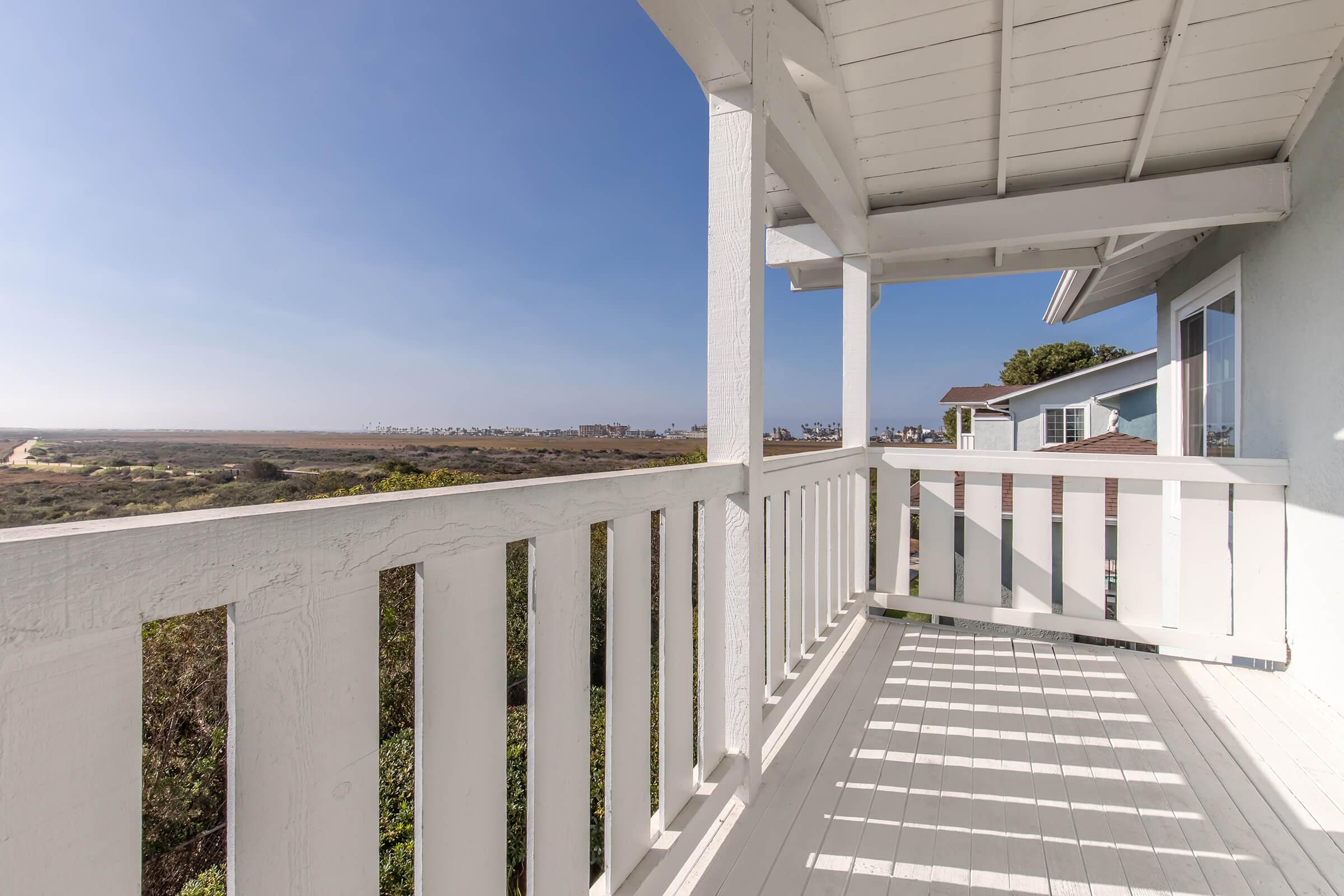 A view from a white wooden balcony overlooking a landscape with rolling hills and a distant coastline. The clear blue sky contrasts with the green vegetation below, providing a serene and inviting atmosphere. The architectural details of the balcony railings are prominent.