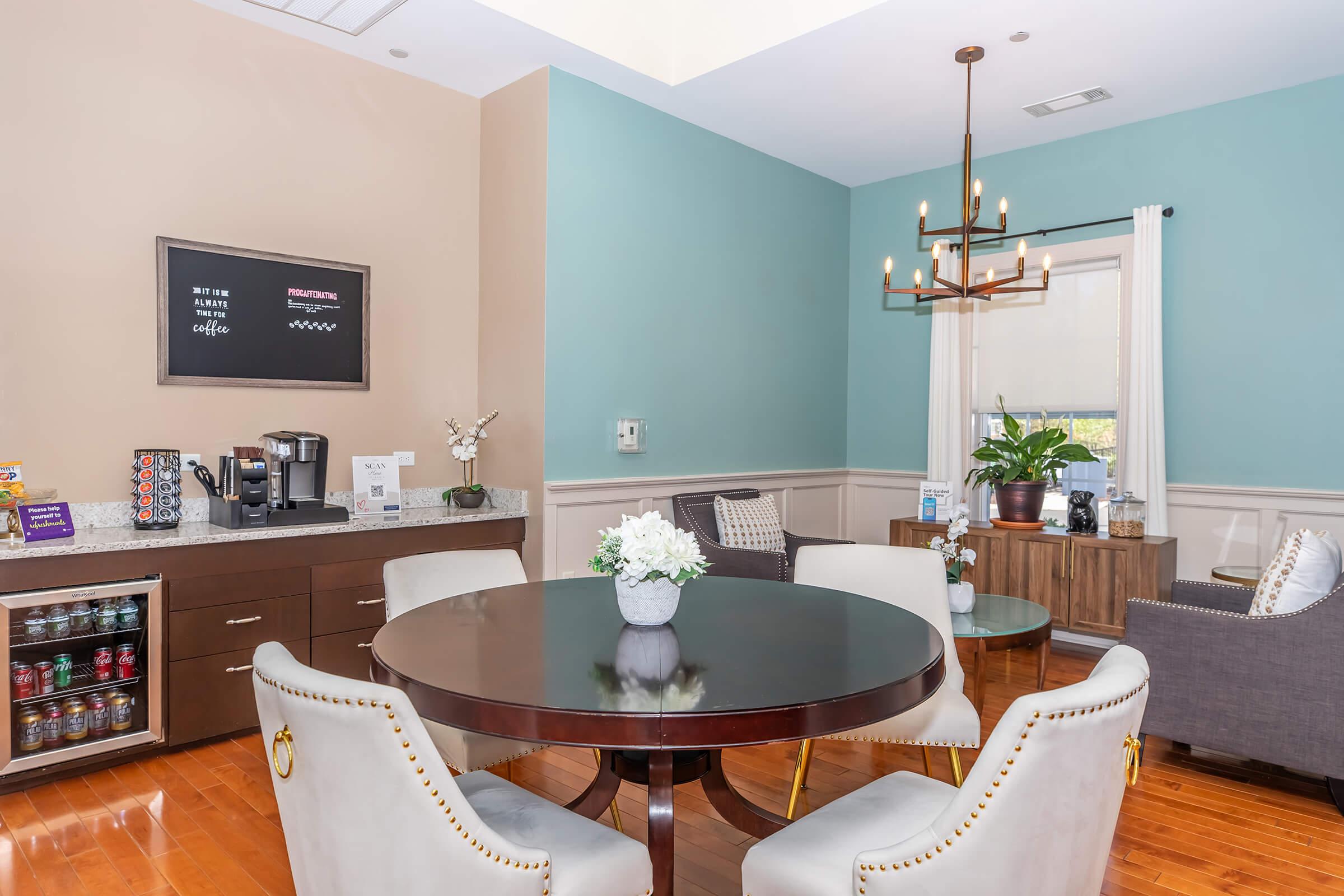 A cozy common area featuring a round dining table with white chairs. There is a coffee machine on a counter with a menu board above it. Two potted plants are visible, along with a side cabinet and a small seating area. The walls are painted in soft beige and blue tones, creating a welcoming atmosphere.