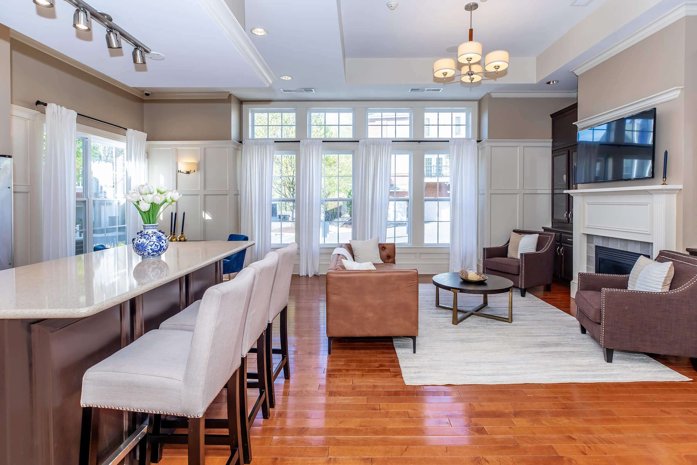Modern living room interior featuring a light color scheme with large windows allowing natural light. There is a stylish bar area with high stools, a seating arrangement with brown chairs, a coffee table, and a decorative vase with tulips. The flooring is hardwood with a light area rug.
