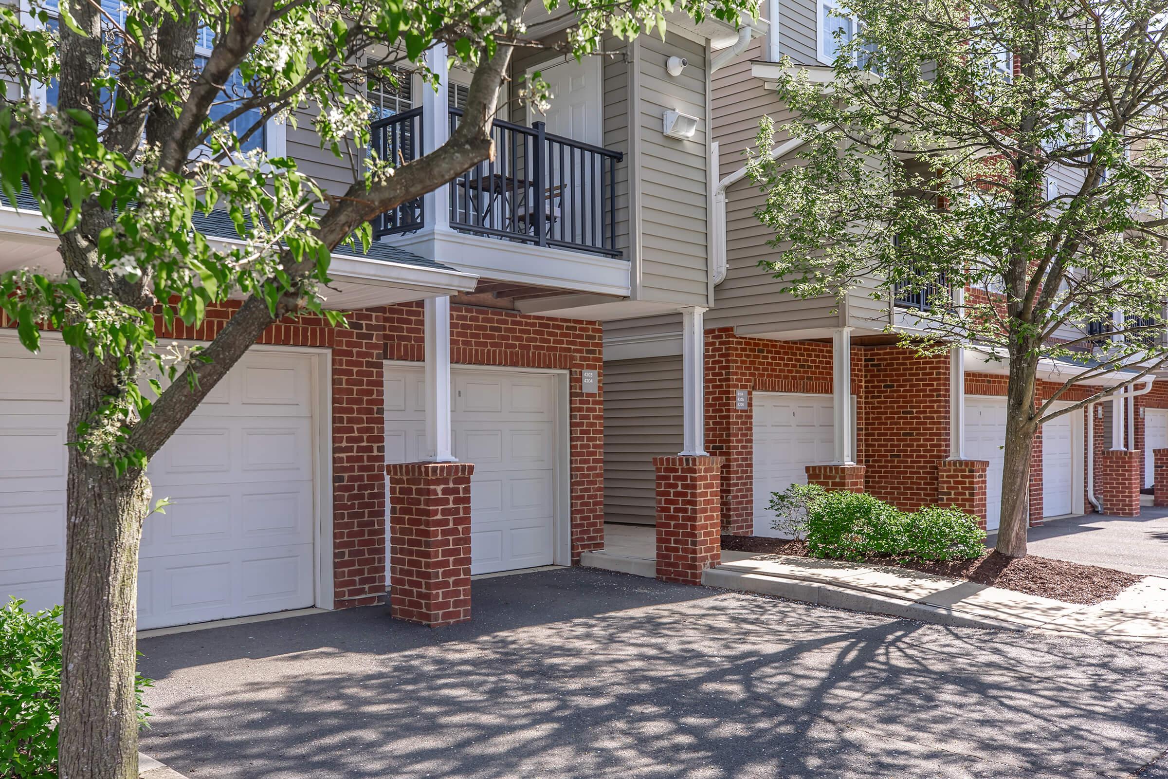 Row of garage doors in a residential area, with brick accents and grey siding on the buildings. Trees provide some shade, and shadows are cast on the pavement. The setting appears neat and well-maintained, ideal for suburban living.
