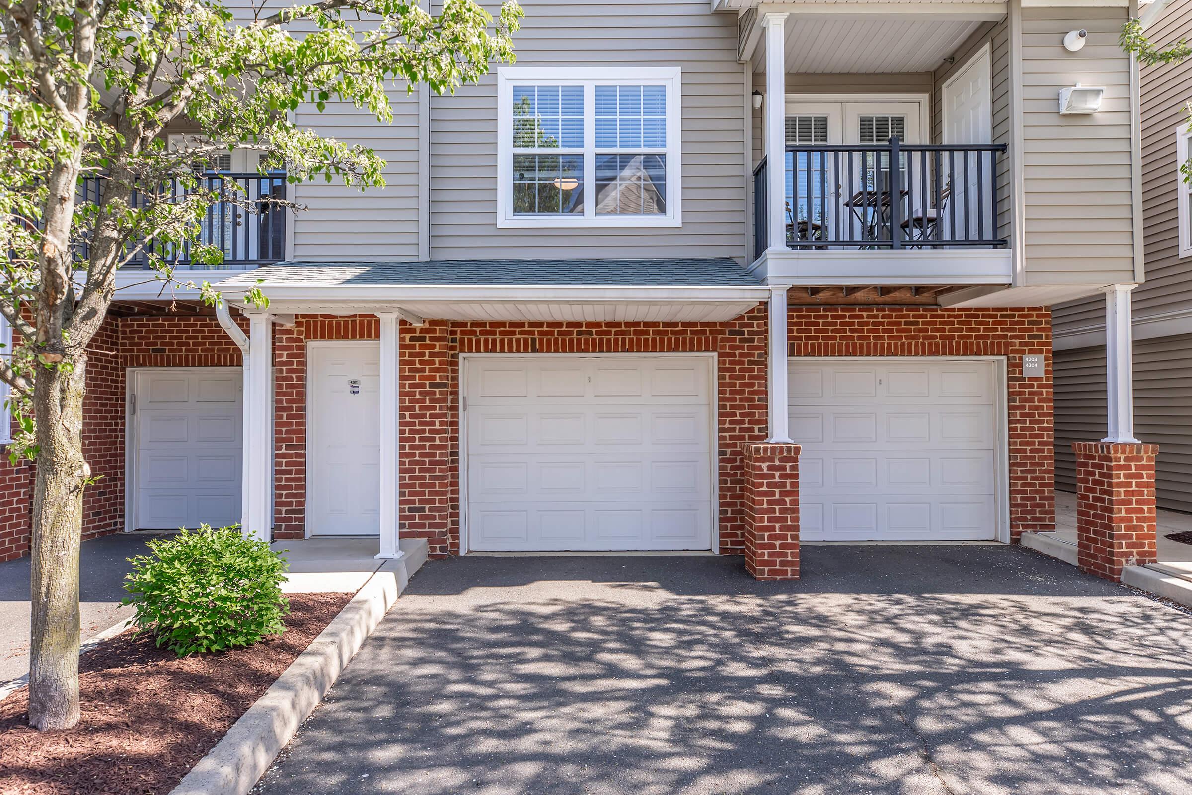A view of a residential building featuring beige siding and two garage doors with white panels. There is a small tree and shrub in front, and a staircase leading to a second-floor balcony with chairs. The driveway is neatly paved with shadows cast by the tree.