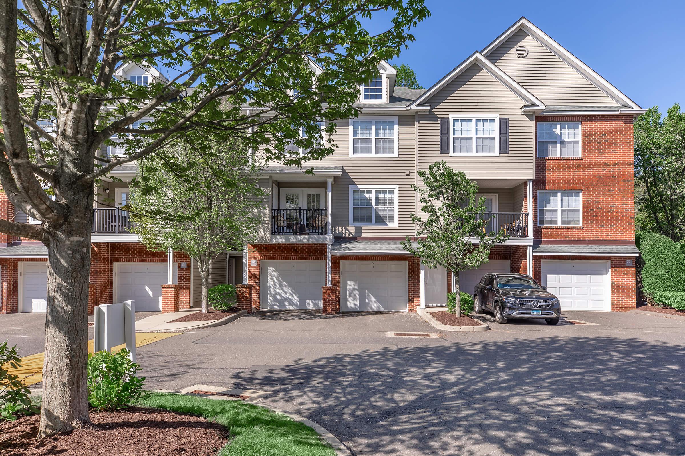 A well-maintained multi-unit residential building with a brick facade and light wood siding. The front features balconies, small trees, and landscaped areas. A black car is parked in front of a garage, and a clear blue sky is visible overhead.