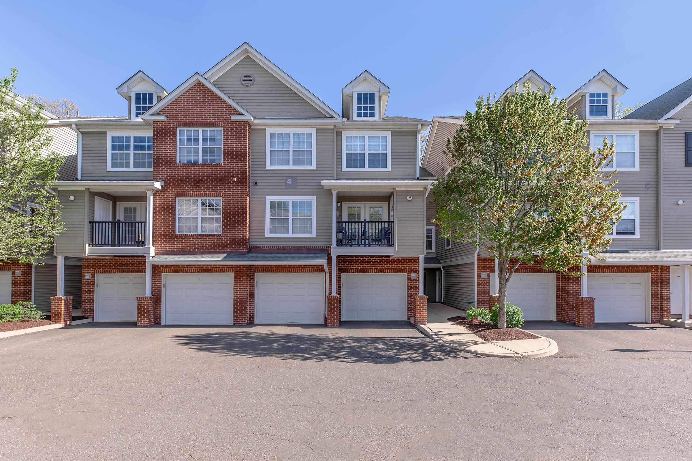 Residential building with multiple units featuring a brick and siding exterior. Each unit has a garage door at the ground level. The front area is landscaped with small trees and shrubs, and the sky is clear and blue. The overall setting appears to be a quiet neighborhood.