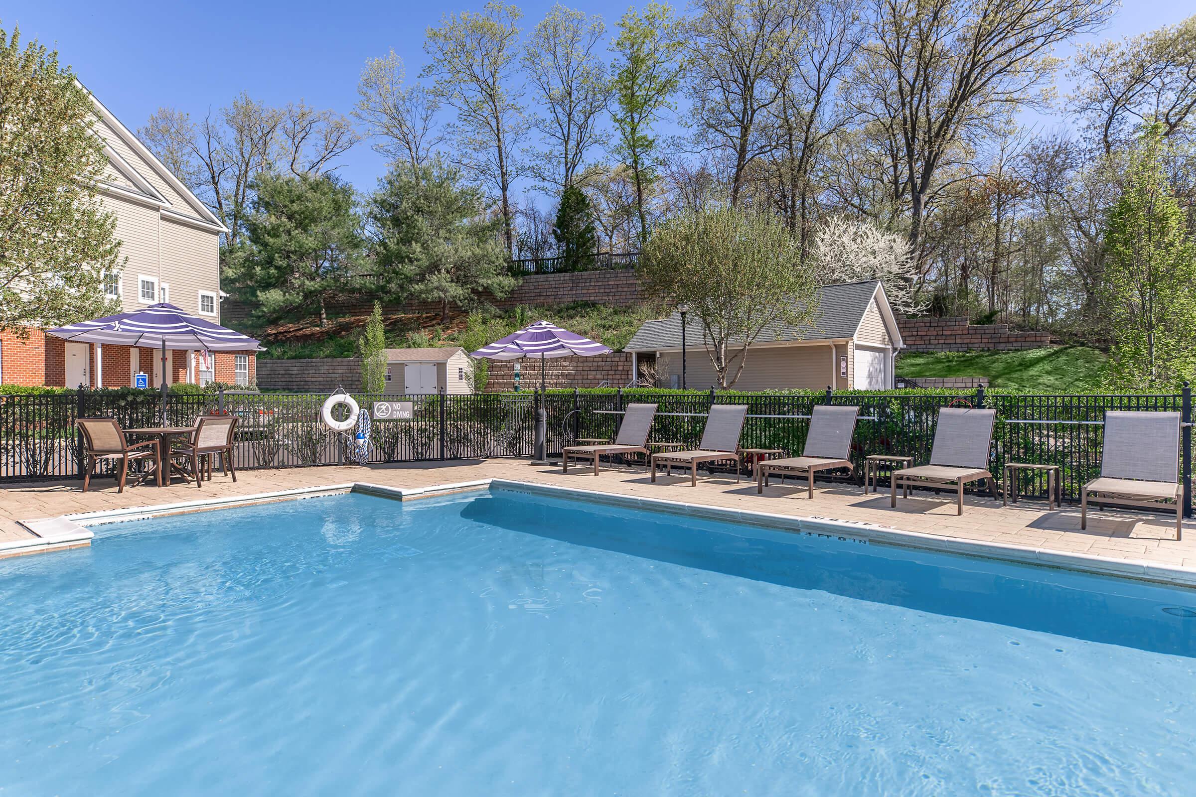 A clear blue swimming pool surrounded by lounge chairs and umbrellas. In the background, there are trees and a building, with a grassy area visible. The scene is set on a sunny day, creating a relaxing outdoor atmosphere.