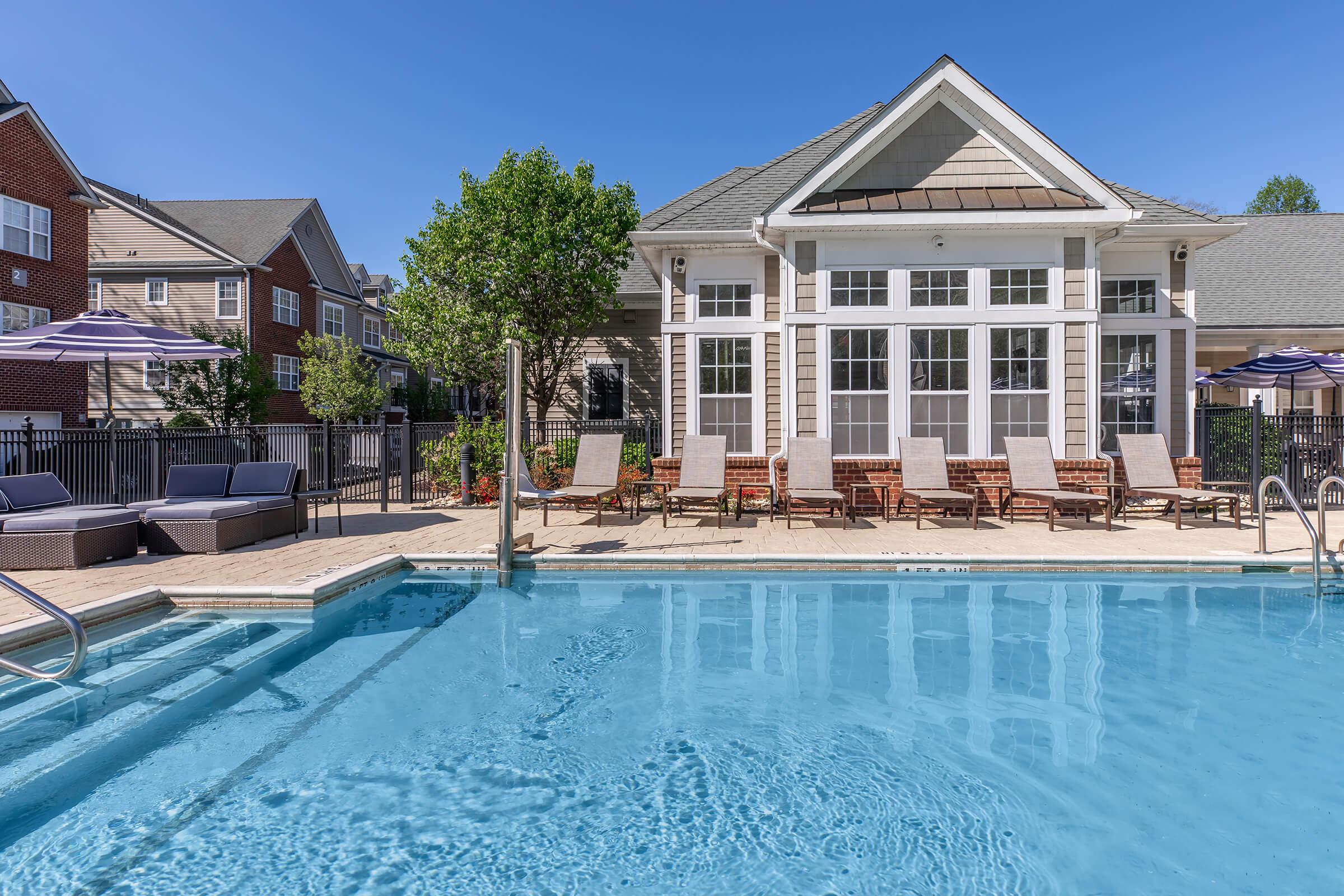 A clear blue swimming pool surrounded by lounge chairs and an elegantly designed building in the background. The area features trees and umbrella-shaded seating, set against a bright blue sky. Ideal for relaxation and outdoor gatherings.