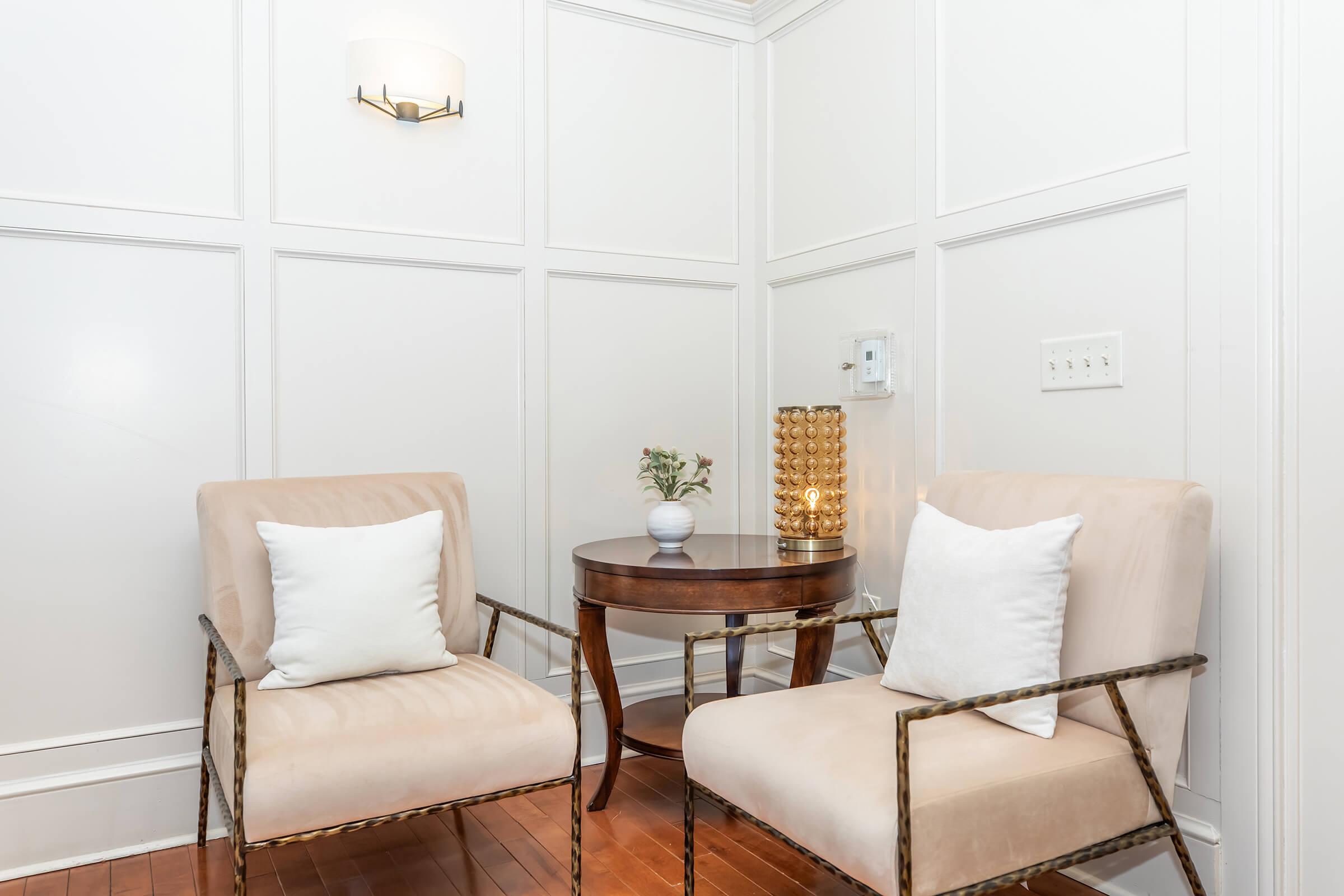 A cozy interior scene featuring two beige armchairs with white throw pillows, positioned next to a small round wooden table. A decorative lamp and a small potted plant are on the table, while the wall behind is paneled with white molding. The warm hardwood floor complements the furnishings.