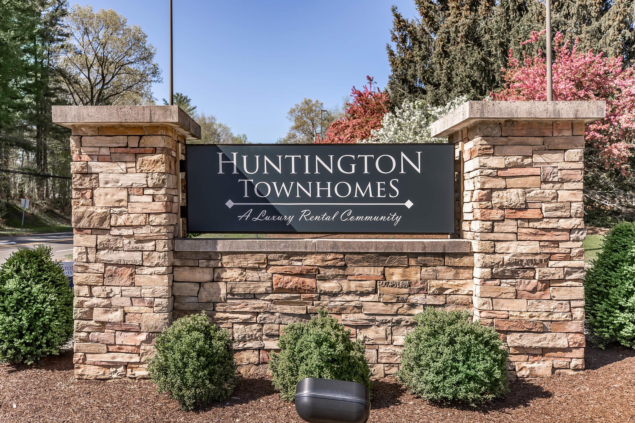 Sign for Huntington Townhomes, featuring the text "Huntington Townhomes" and "A Luxury Rental Community," set against a backdrop of landscaping with green shrubs and trees. The sign has a stone base and a black panel for the text.