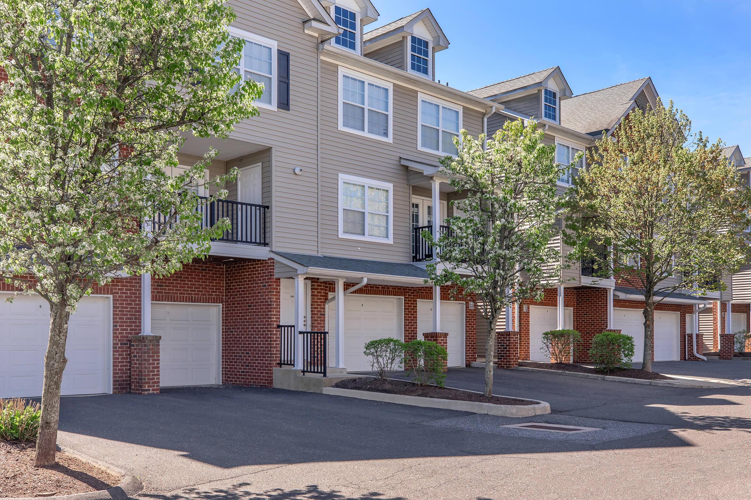 A row of modern townhouses with gray siding and brick accents. Each unit features a garage door, a small porch, and balconies. The landscaping includes small trees and shrubs, and the scene is set under a clear blue sky, suggesting a sunny day.