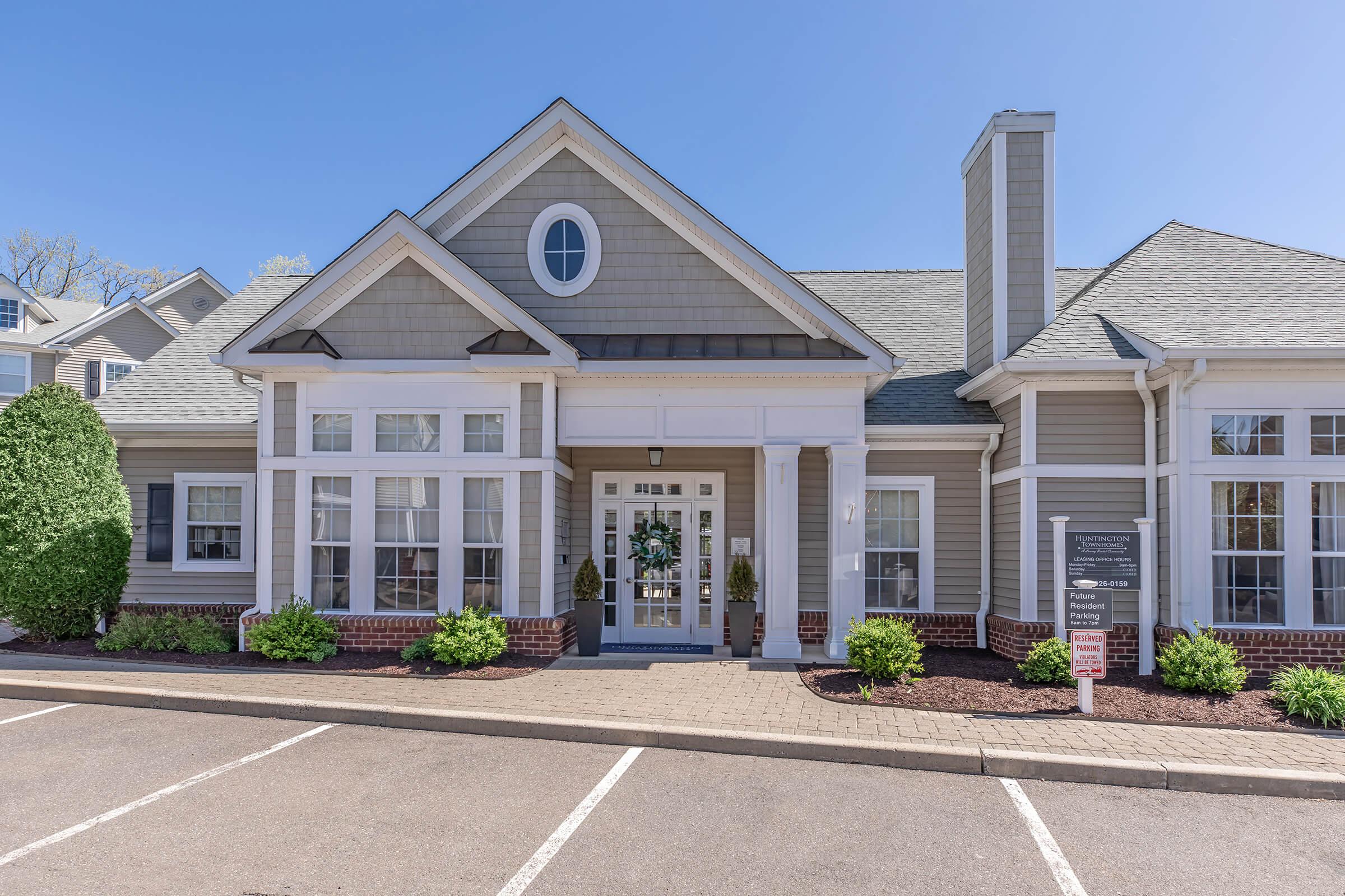 Exterior view of a modern, light-colored building featuring a well-maintained entrance with large windows, a decorative door, and neatly trimmed shrubs. The parking area in front is paved with clear white lines indicating parking spots, and the sky is bright and blue, suggesting a sunny day.
