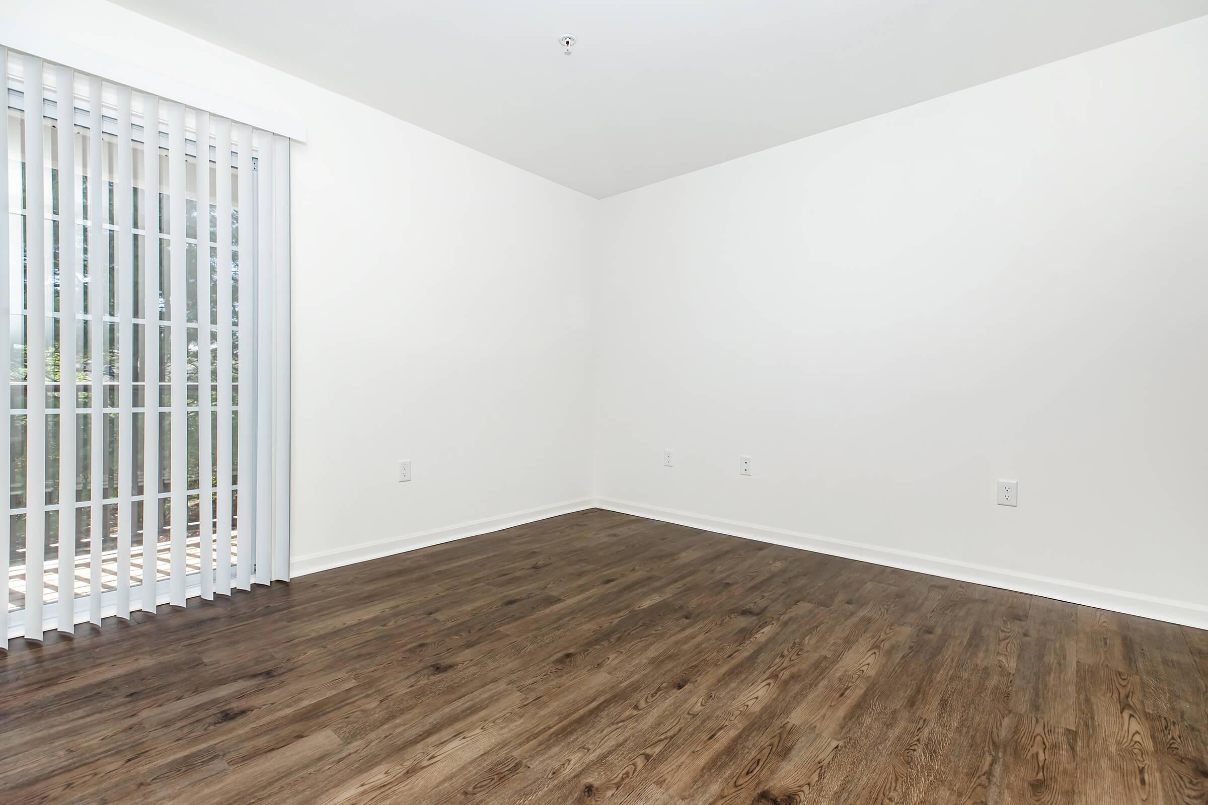 Empty room with light-colored walls and dark wooden flooring. A large window with vertical blinds allows natural light to enter, showcasing the spaciousness of the area. The room is unfurnished, emphasizing its clean and minimalistic design. No decorations or furniture are present.