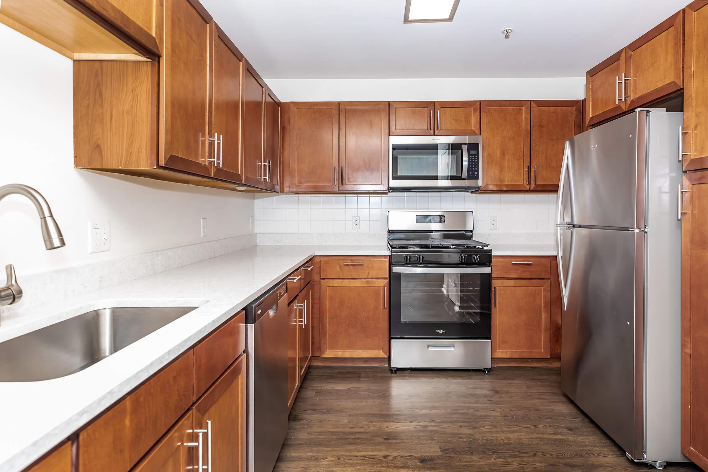 A modern kitchen featuring wooden cabinetry, a stainless steel refrigerator, and a gas stove with an oven. The countertop is made of light-colored stone, and there is a sleek sink with a faucet. The kitchen has an open layout with ample storage space and a clean, contemporary design.