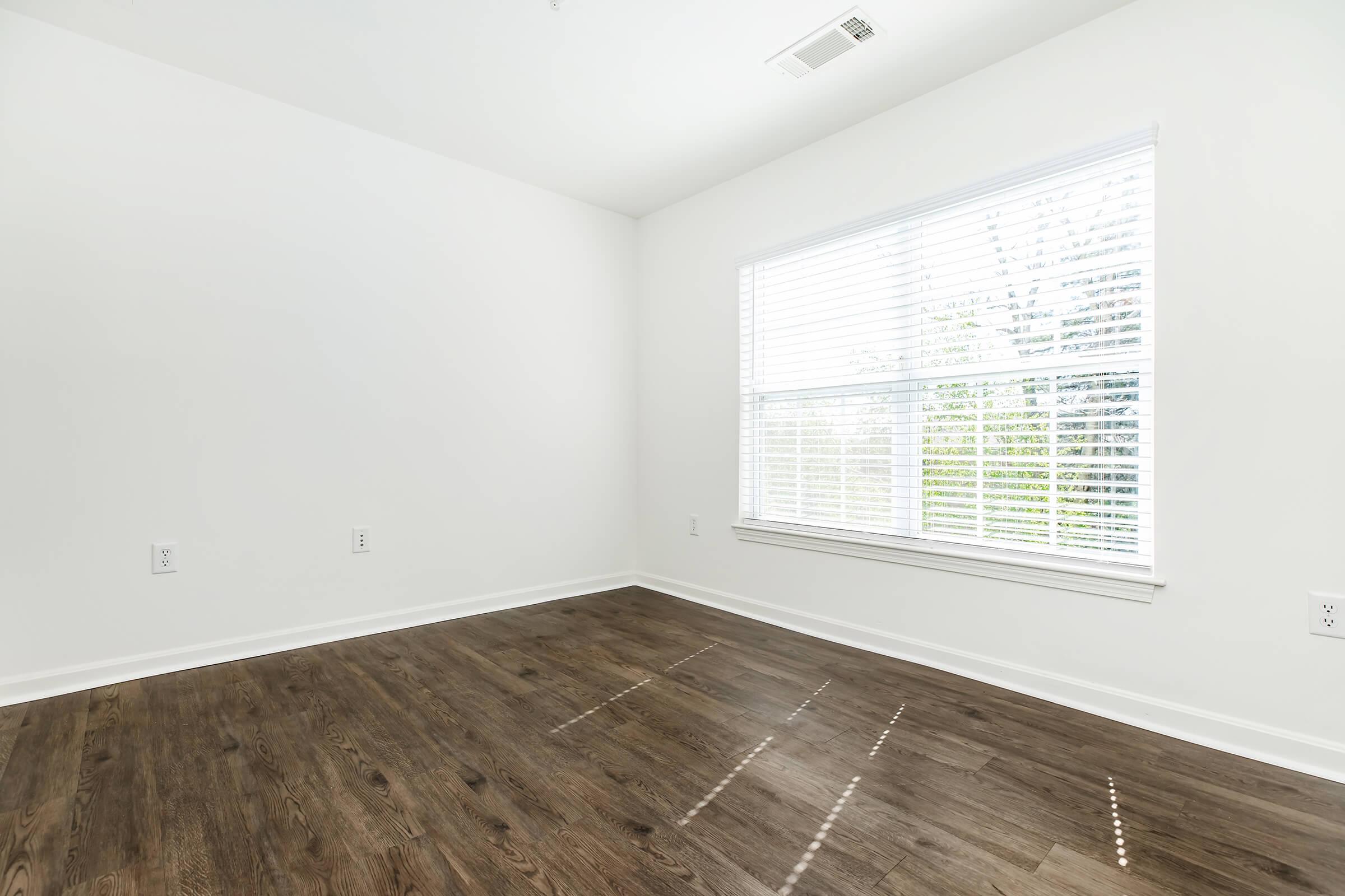 A bright, empty room with white walls and large window covered by white blinds. Natural light shines in, highlighting the wooden floor. The room is unfurnished, creating a spacious and open atmosphere.