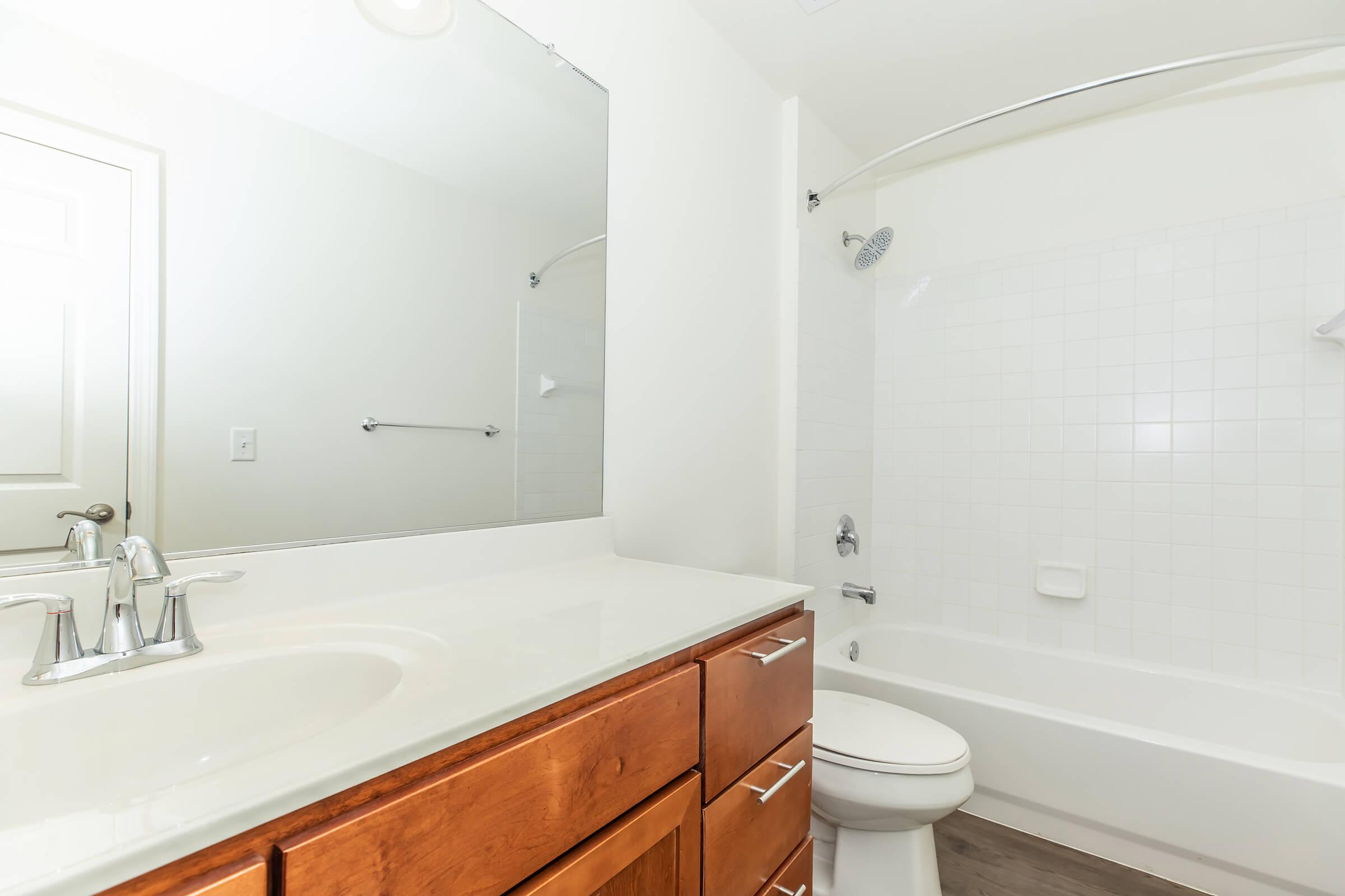 A modern bathroom featuring a white tiled shower with a glass shower rod, a white bathtub, a sleek vanity with wooden cabinets, a sink with chrome faucet, and a large mirror above the sink. The bright space has neutral-colored walls and a light wooden floor.