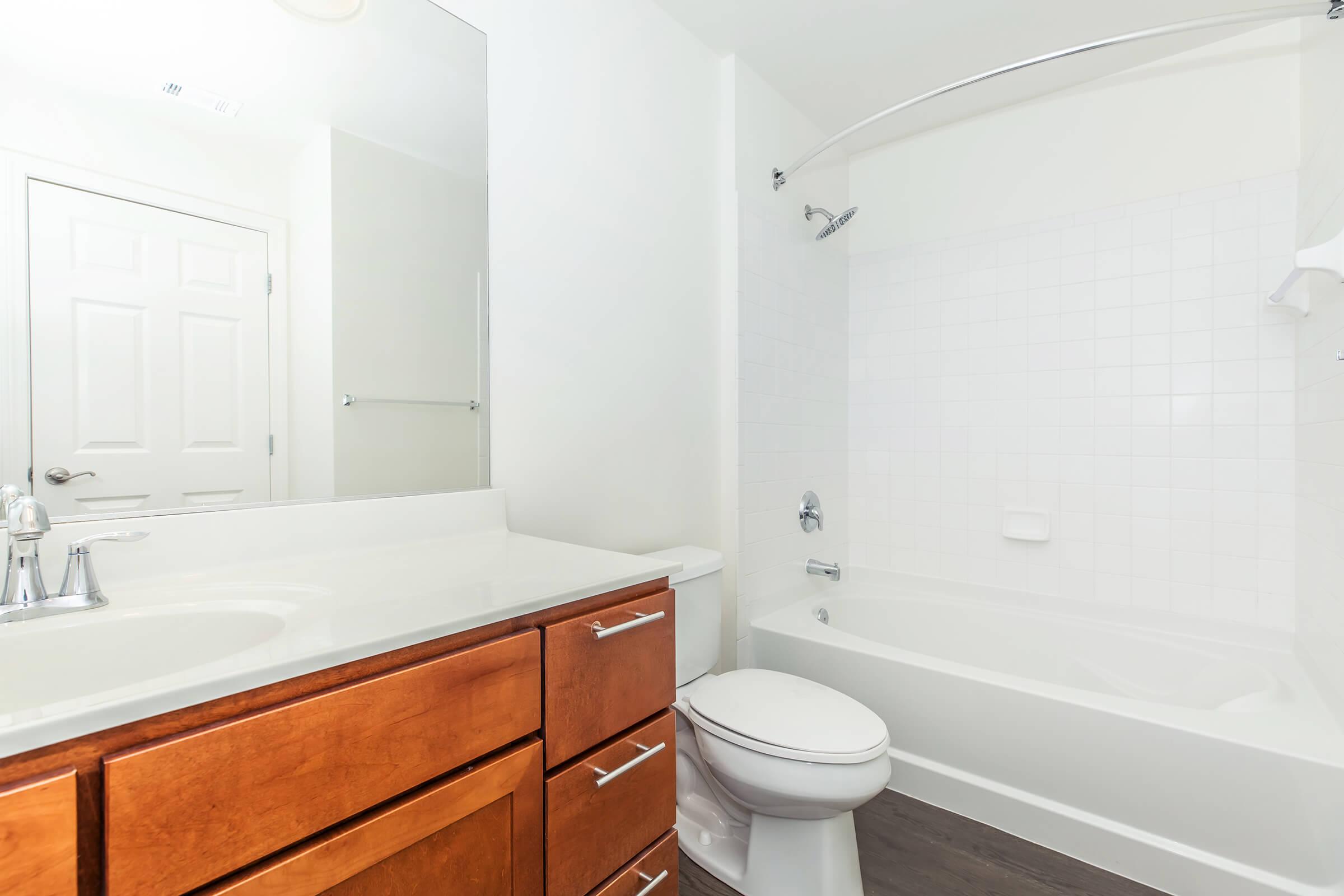 A clean and modern bathroom featuring a bathtub with a shower, a white tiled wall, a large mirror above a dark wood vanity with storage drawers, and a toilet. The overall design emphasizes bright lighting and a spacious feel, with a door visible in the background.