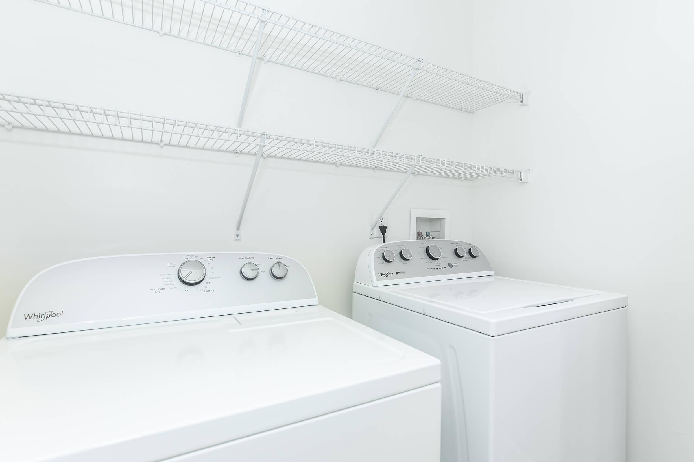 A clean laundry room featuring two white Whirlpool appliances: a front-loading washing machine on the left and a top-loading dryer on the right. Above the appliances, there are two wire shelves for storage. The walls are painted white, creating a bright and spacious atmosphere.