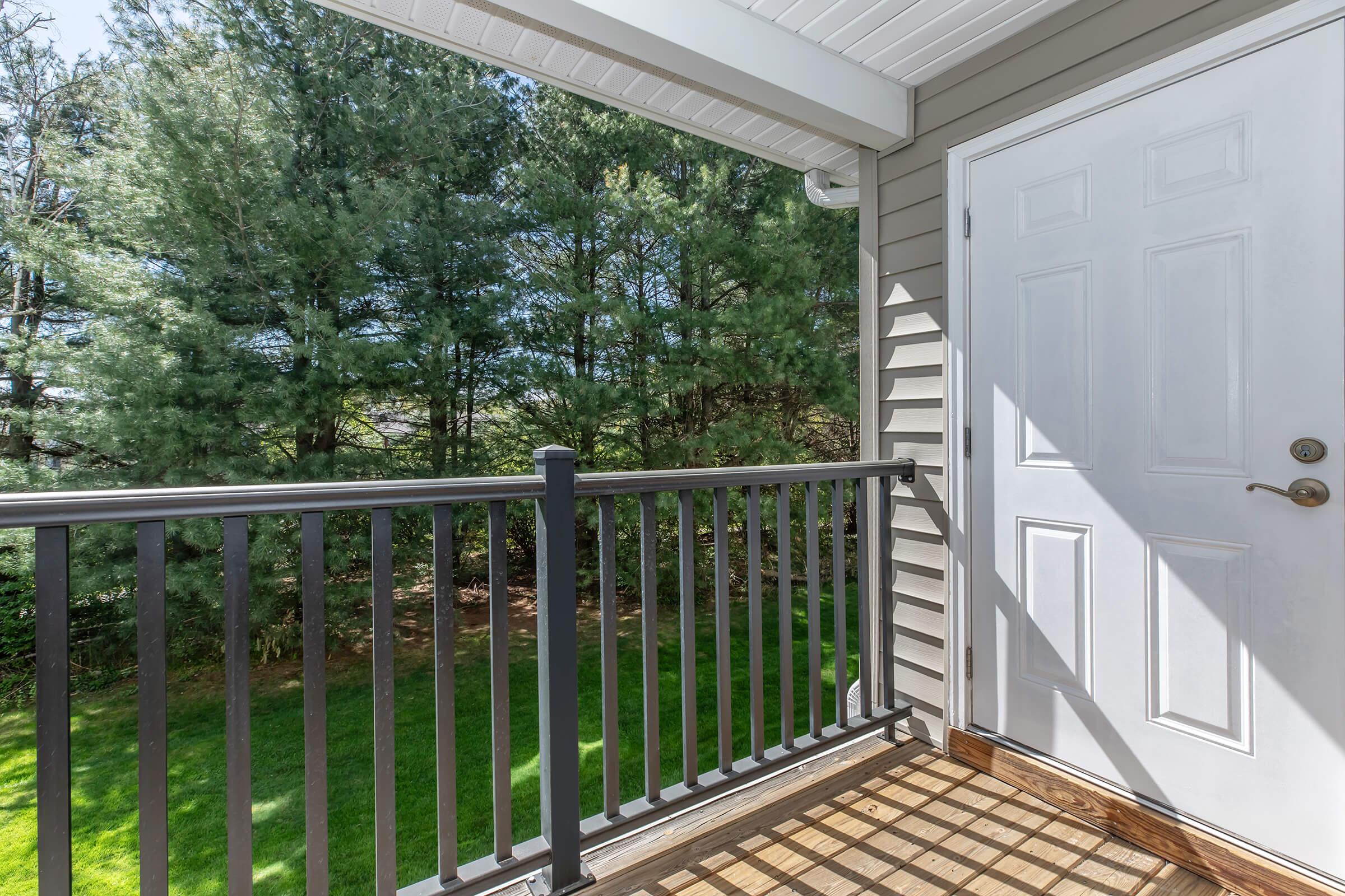 A balcony view featuring a white door and black railing, overlooking a grassy area surrounded by tall green trees on a sunny day. The floor is wooden with a natural finish, and sunlight casts shadows on the balcony floor.