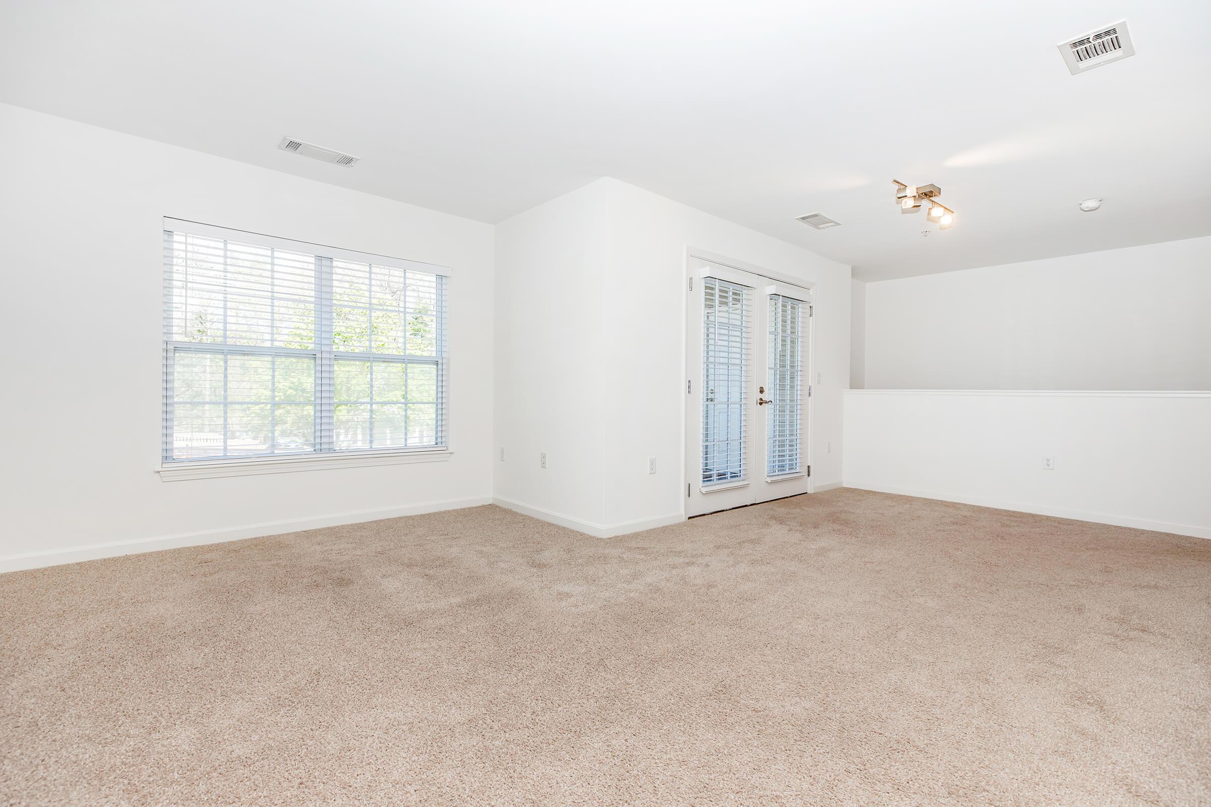 A bright, empty room with light beige carpet, white walls, and large windows allowing natural light. There are double doors leading outside, and a ceiling light fixture. The space is unadorned, offering a blank canvas for potential furnishings and decor.