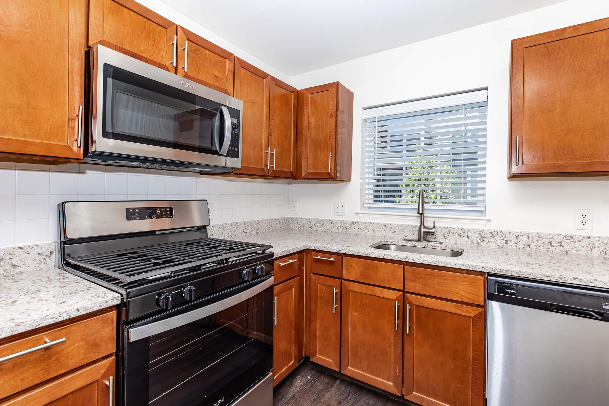 Modern kitchen featuring wooden cabinets, a stainless steel microwave, a gas stove, and a sleek dishwasher. The countertop is made of light-colored granite, and there's a window with blinds providing natural light. The overall design is clean and contemporary.