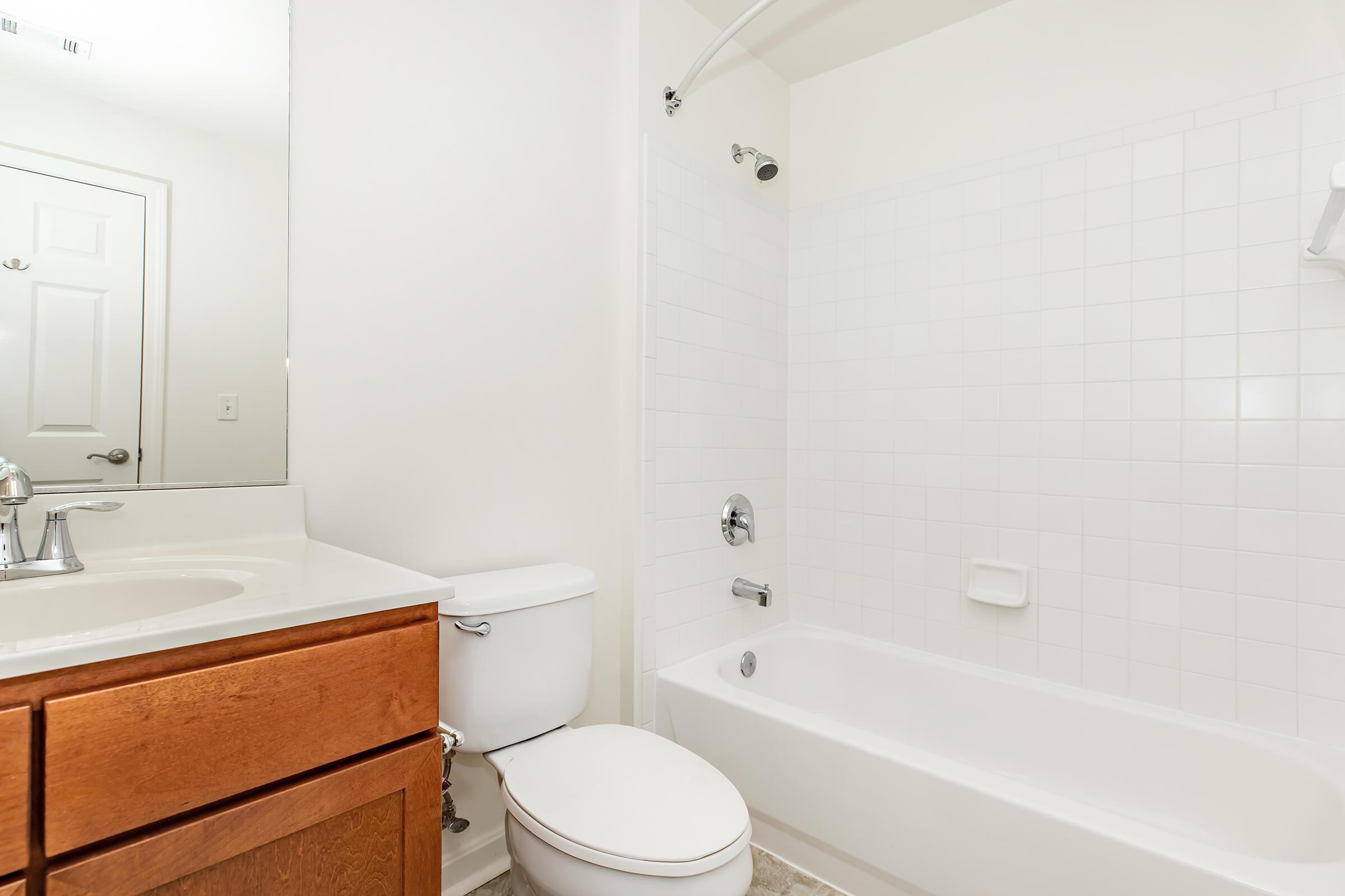A clean and modern bathroom featuring a white tiled shower and bathtub combination, a single sink vanity with wooden cabinetry, a toilet, and a large mirror above the sink. The overall color palette is light and neutral, creating a spacious and inviting atmosphere.