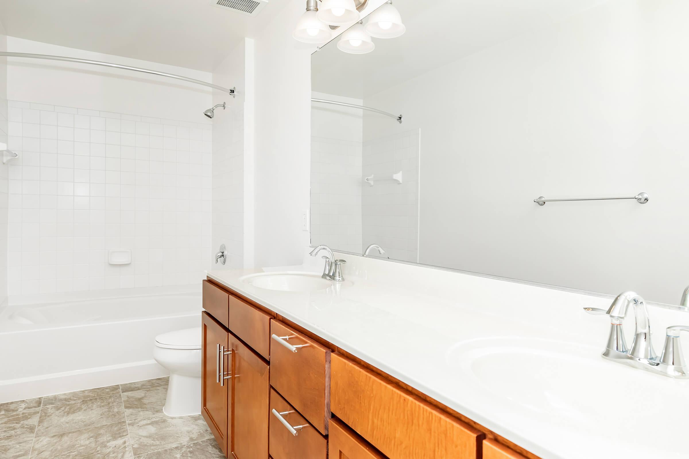 A clean, modern bathroom featuring a double sink vanity with wooden cabinets, a large mirror, a white bathtub, and a separate shower area. The walls are light-colored and the flooring is a subtle tile pattern, creating a bright and spacious feel.