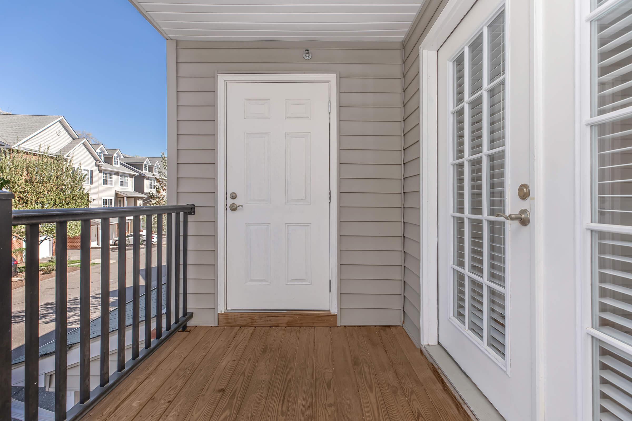 A covered porch featuring a white door and glass-panel double doors, with a wooden floor. The exterior walls are gray with horizontal siding, and the area is sunny, showcasing a neighborhood view in the background with other buildings and greenery visible.