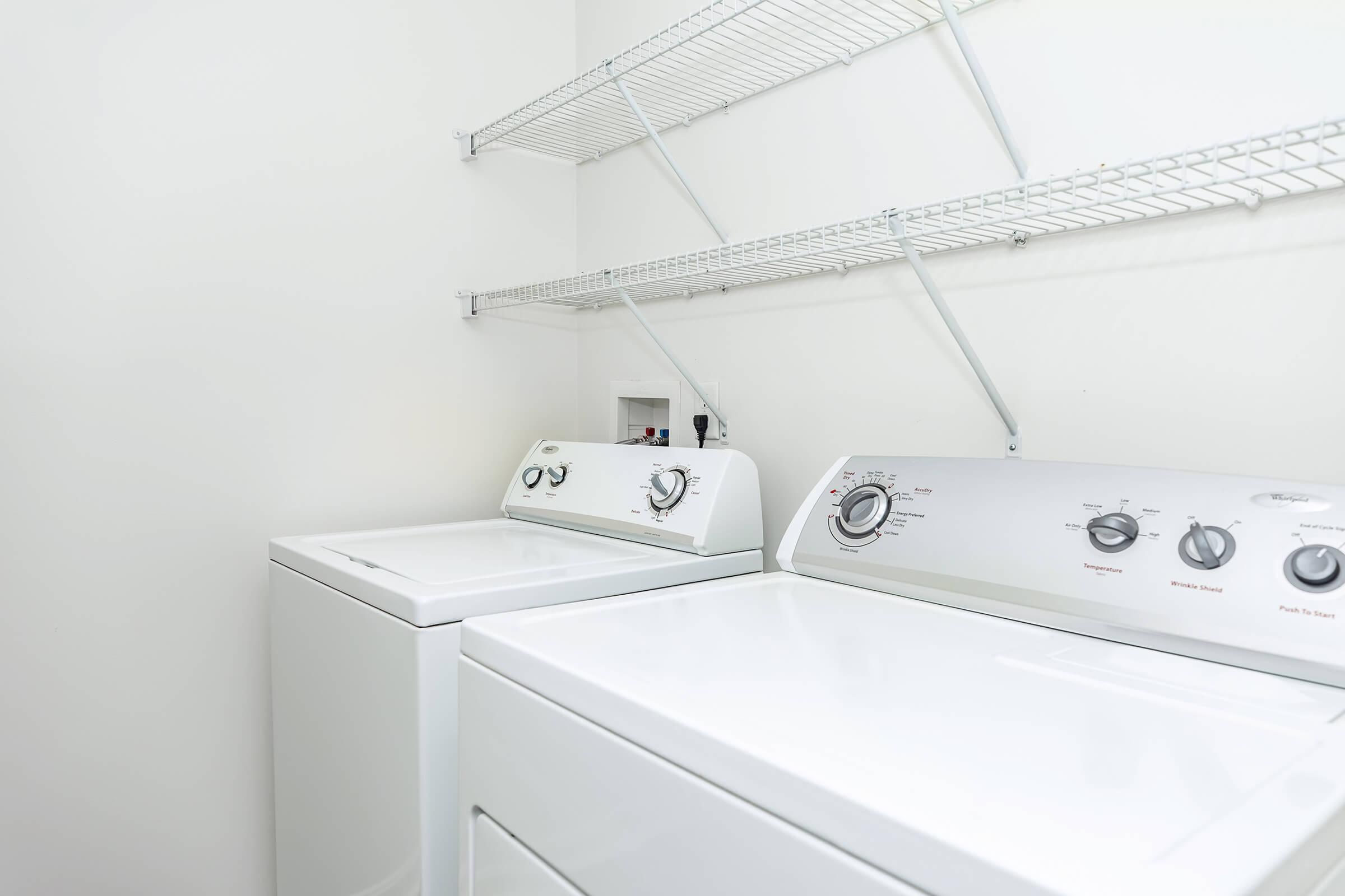 A clean laundry room featuring a white washing machine and dryer side by side, with control knobs visible. Above them, there are metal shelving units mounted on the wall, providing storage space. The walls are painted white, creating a tidy and bright environment.
