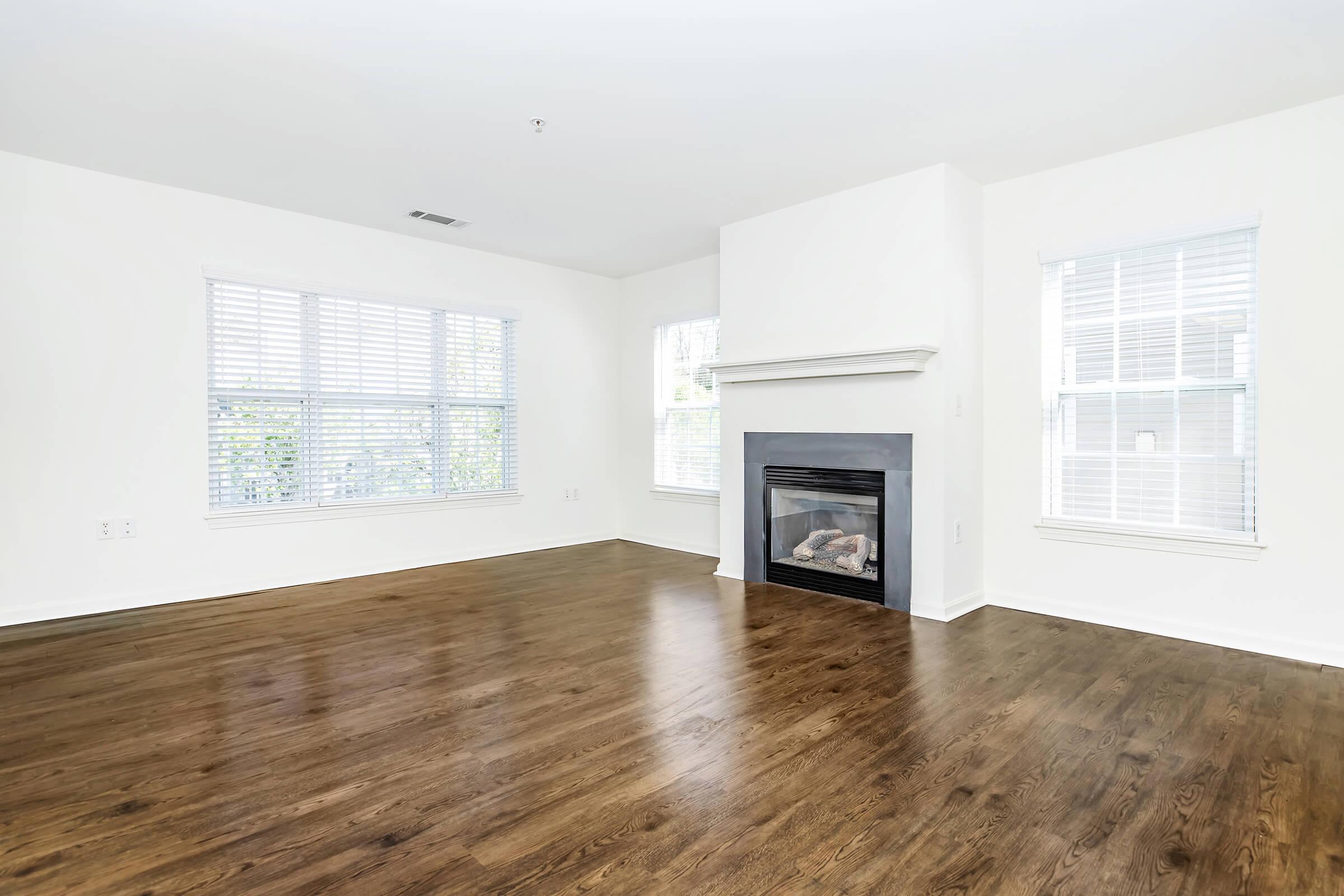 A bright, spacious living room with hardwood floors, large windows allowing natural light, and a white fireplace on the wall. The walls are painted white, creating a clean and open atmosphere.