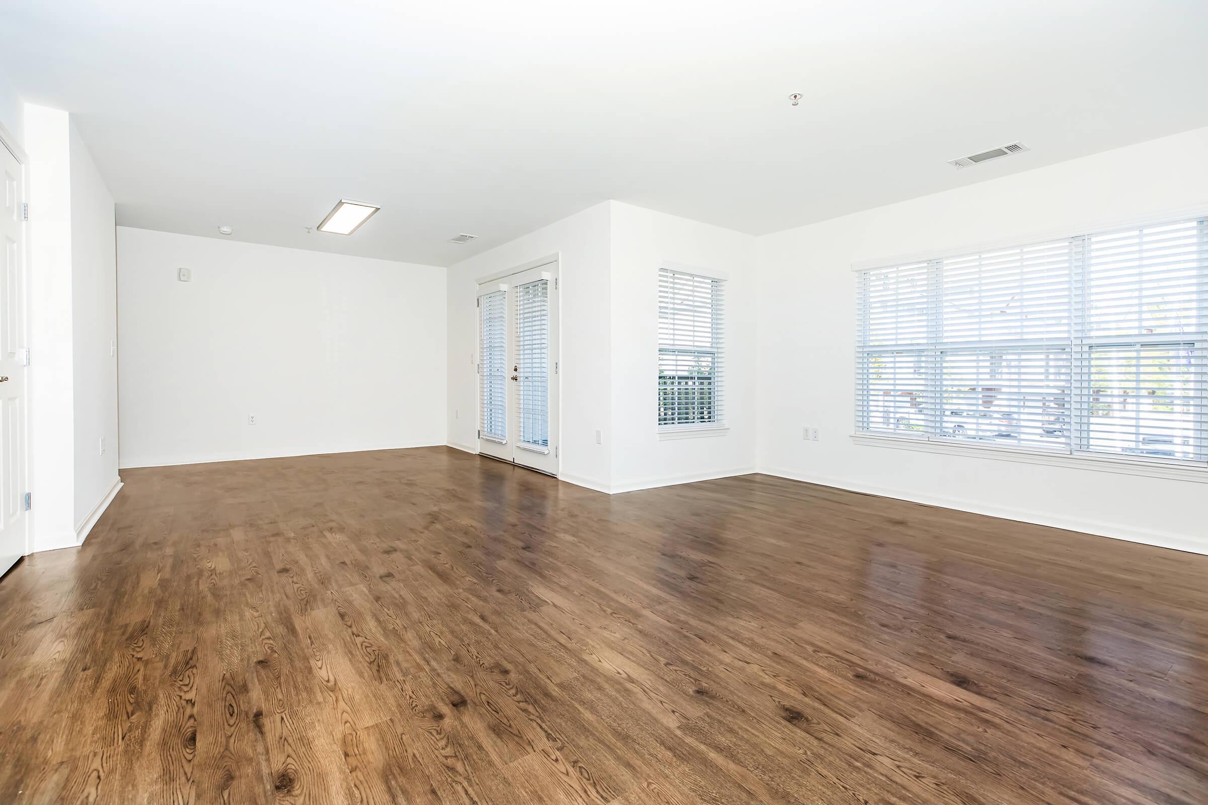 A spacious, empty room with light-colored walls and hardwood flooring. Large windows with white blinds allow natural light to enter, and there are sliding glass doors leading to an outdoor space. The room has a bright, airy feel, ideal for various interior design possibilities.