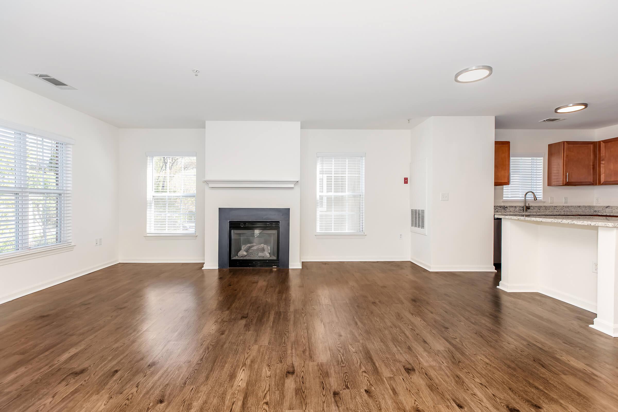 A spacious living room with hardwood flooring and a white fireplace as the focal point. Large windows allow natural light, and the open layout connects to a nearby kitchen featuring wooden cabinets. The walls are painted white, creating a bright and inviting atmosphere.