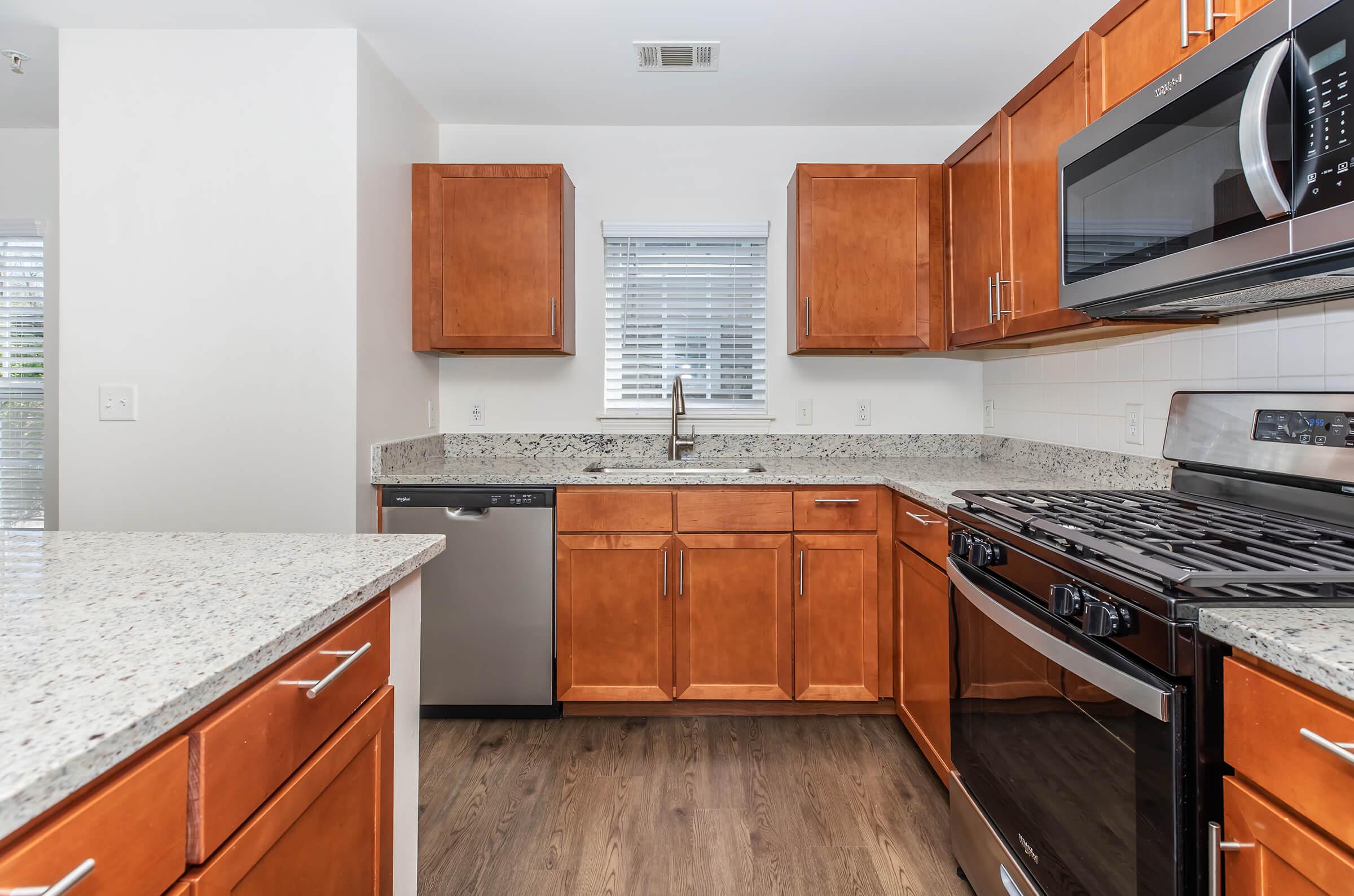 Modern kitchen featuring warm wood cabinetry, a granite countertop, stainless steel appliances including a microwave and a dishwasher, and a gas stove. Natural light enters through a nearby window, illuminating the space. The design is clean and functional, with a contemporary feel.