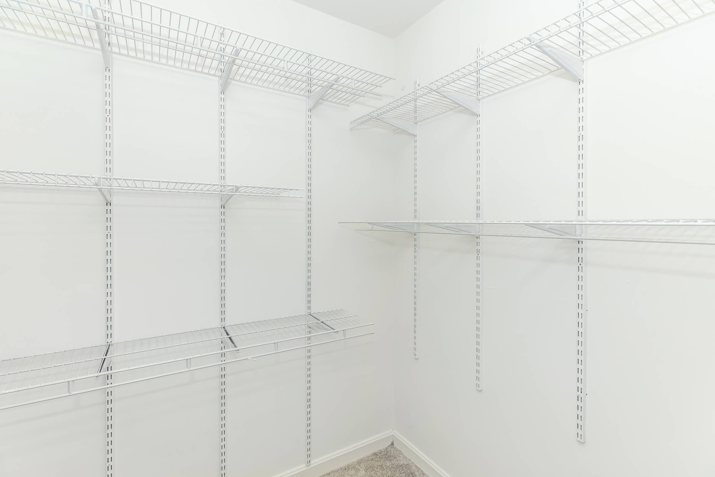 Empty closet with white walls and wire shelving. The shelves are mounted on the walls, providing storage space. The floor is carpeted, and the overall appearance is clean and organized, awaiting items to be stored.