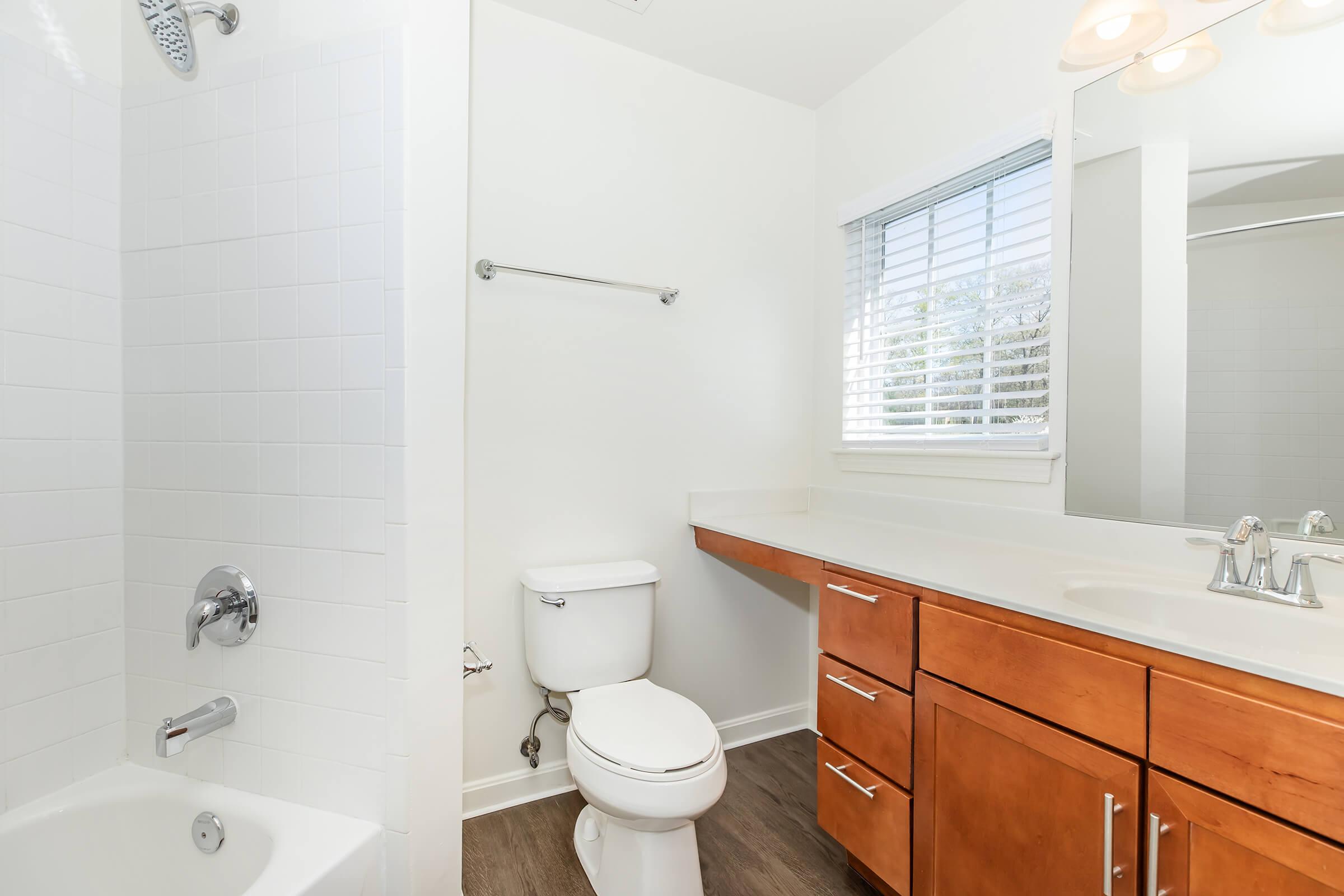Modern bathroom featuring a white tiled shower, a toilet, and a double-sink vanity with wooden cabinets. Natural light enters through a window with horizontal blinds, illuminating the clean, minimalist design. The floor is a neutral tone, complementing the overall aesthetic.