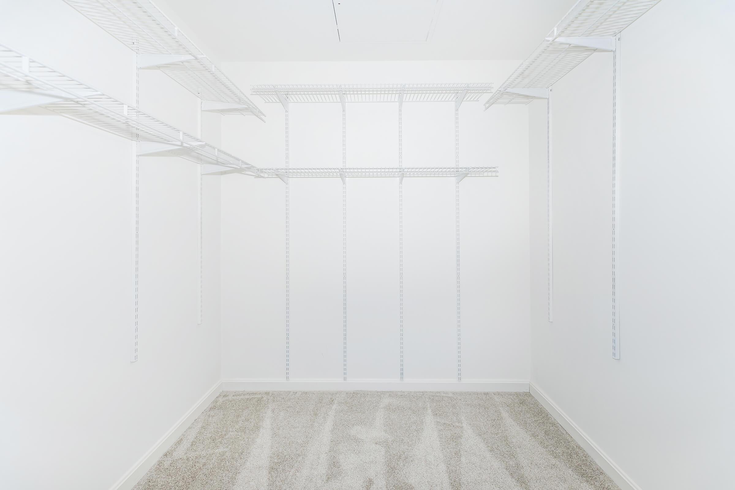 An empty walk-in closet featuring white wire shelving units against plain white walls. The floor is carpeted in light gray, creating a clean and spacious appearance. The shelves are positioned at various heights, providing opportunities for organization and storage.