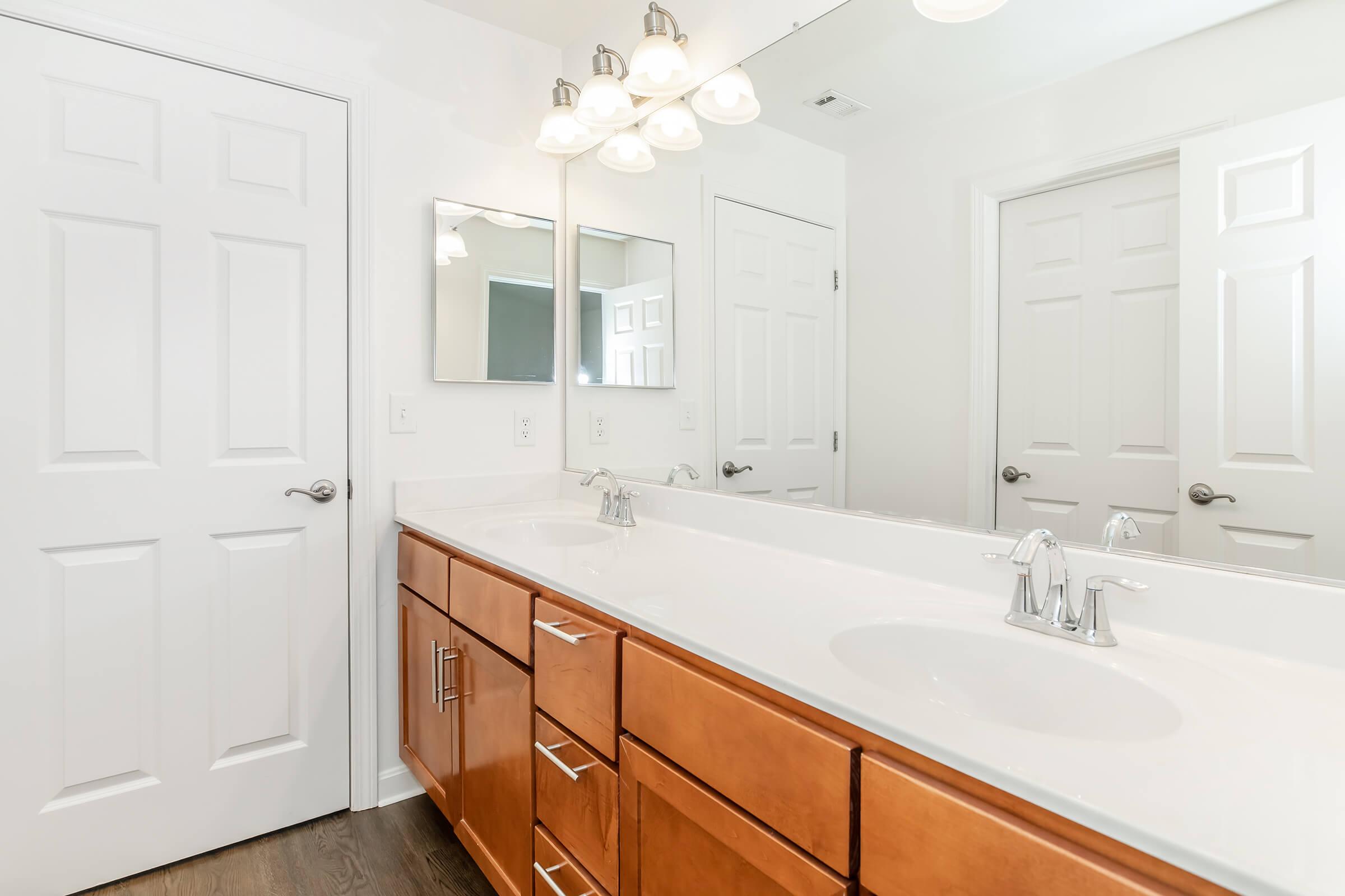 A modern bathroom featuring a double vanity with a white countertop and two sinks, wooden cabinets below, and a large mirror above. The walls are painted white, and there are two doors visible, possibly leading to other rooms. Soft lighting fixtures hang above the mirror.