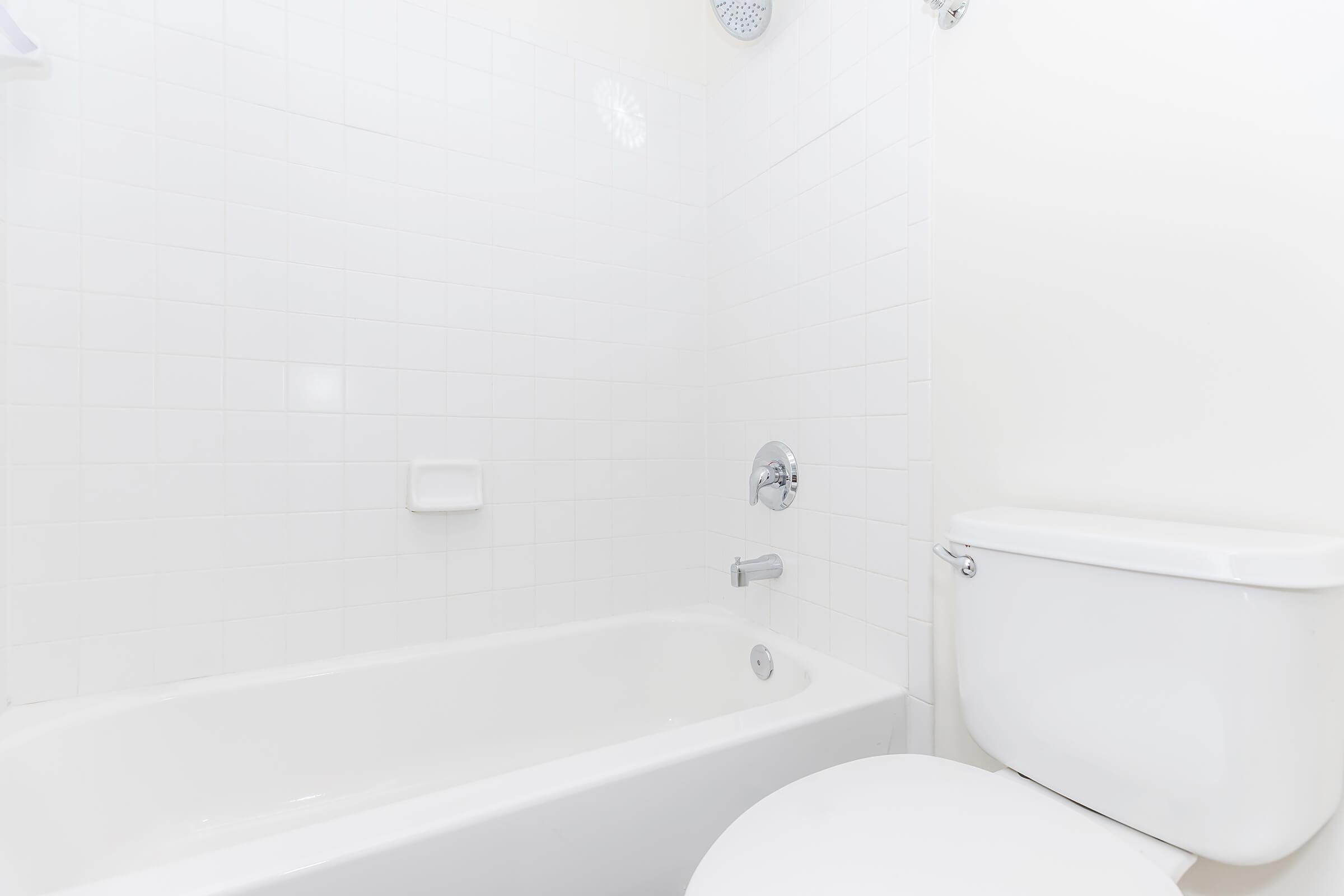 A clean, modern bathroom featuring a white bathtub, a wall-mounted showerhead, and a white toilet. The walls are tiled with smooth, white ceramic tiles, creating a bright and minimalist aesthetic. Natural light enhances the fresh and uncluttered look of the space.