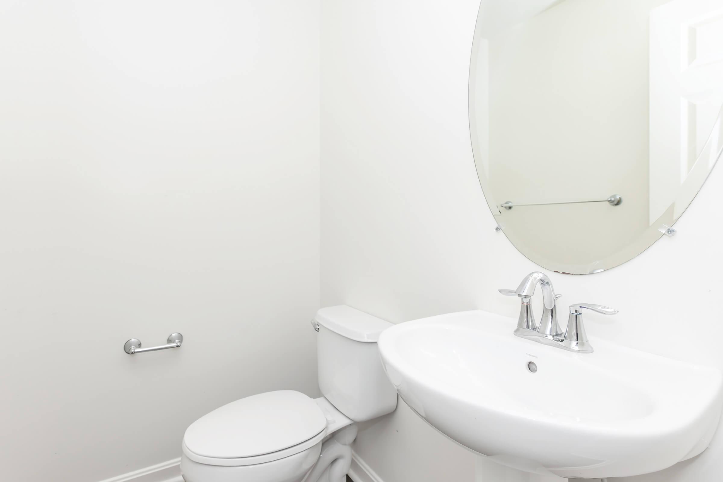A clean and minimalist bathroom featuring a white toilet, a round mirror above a white sink with two silver faucets, and a towel rack on the wall. The walls are plain white, creating a bright and airy atmosphere.