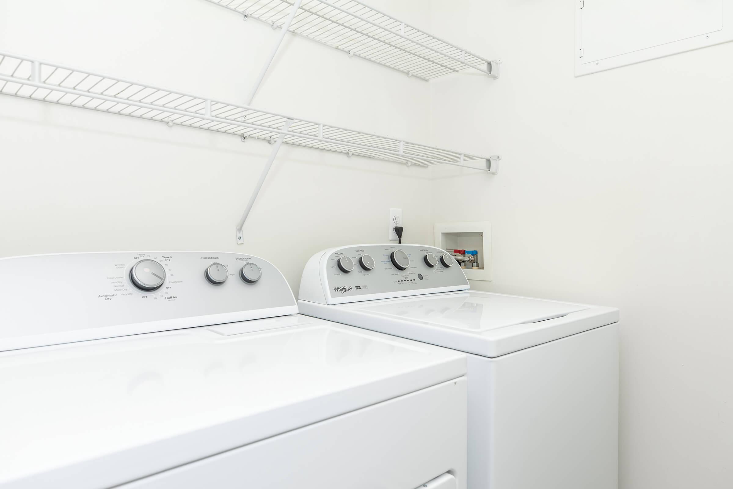 A clean laundry room featuring two white washing machines side by side on a light-colored wall. Above, there is a metal wire shelf for storage. The space is bright and uncluttered, creating an inviting and functional environment for laundry tasks.