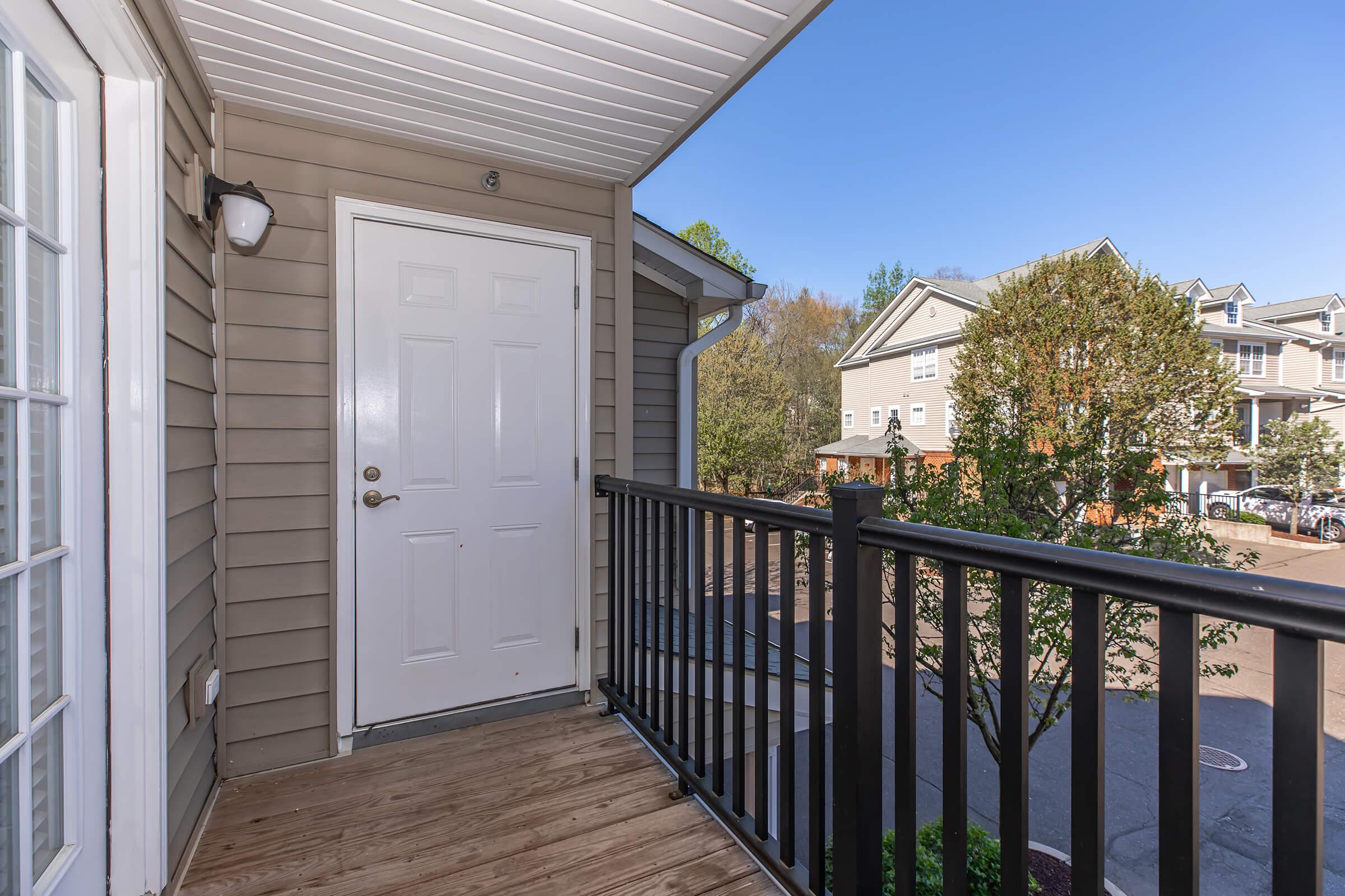 View of a balcony with a wooden floor and black railing, featuring a closed white door. In the background, there are trees and multiple residential buildings under a clear blue sky. The scene suggests a quiet suburban environment.