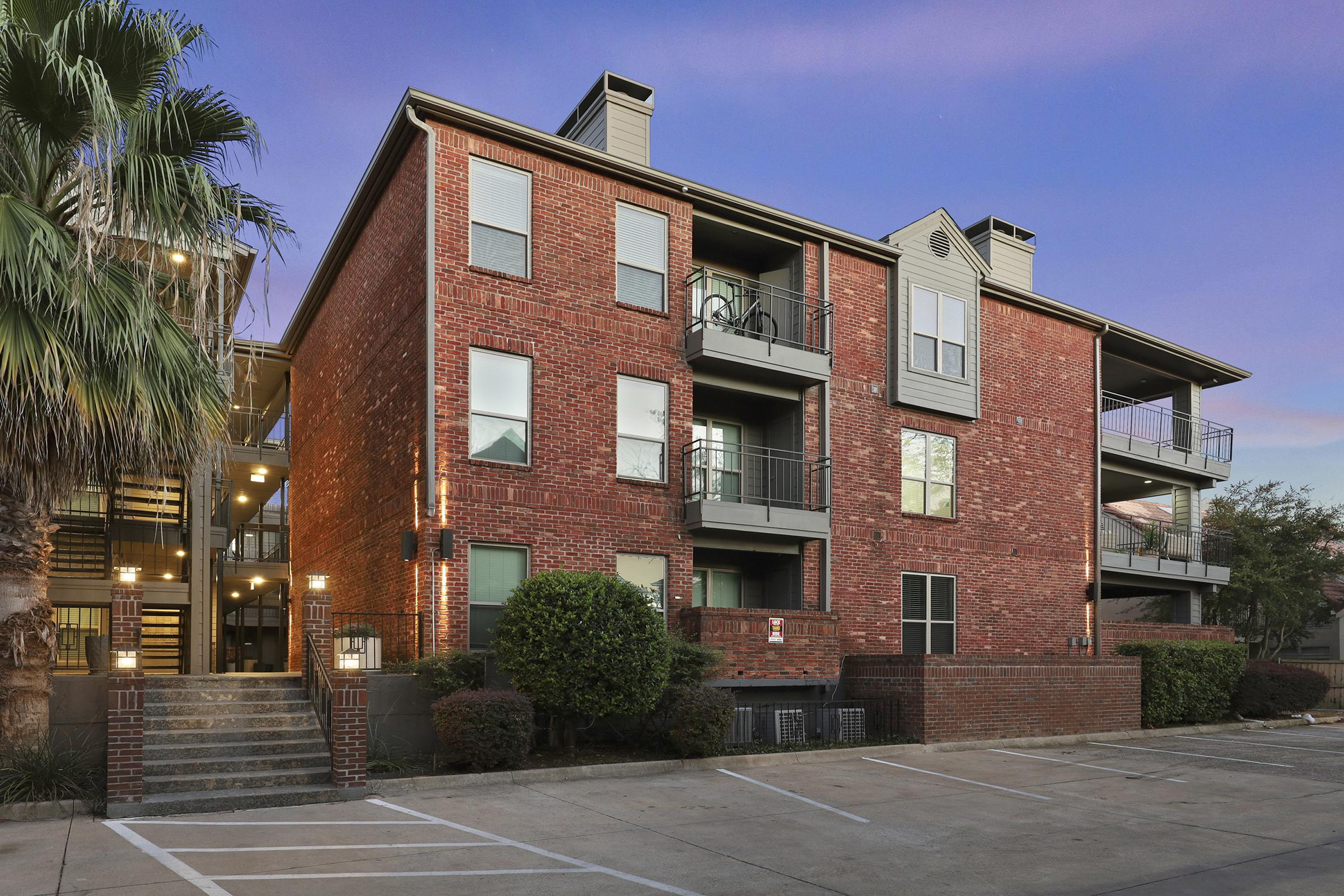 Three-story brick apartment building with balconies, surrounded by palm trees and parking spaces. The entrance features a staircase leading to the lobby, and the building is bathed in warm evening light against a twilight sky.