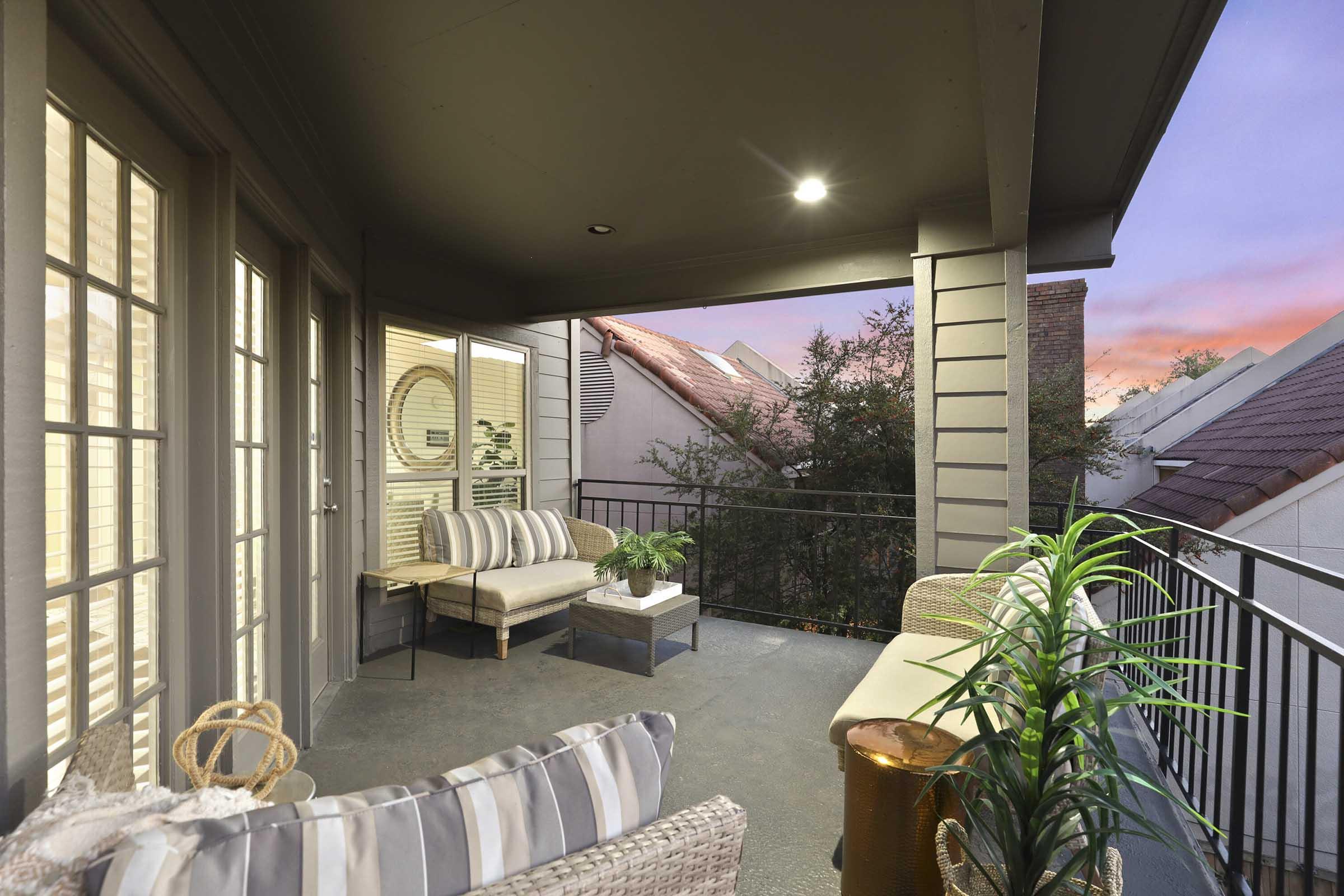 Cozy balcony area featuring a comfortable seating arrangement with striped cushions, a small side table, and greenery plants. The setting offers a peaceful atmosphere with a view of rooftops, illuminated by soft evening light.