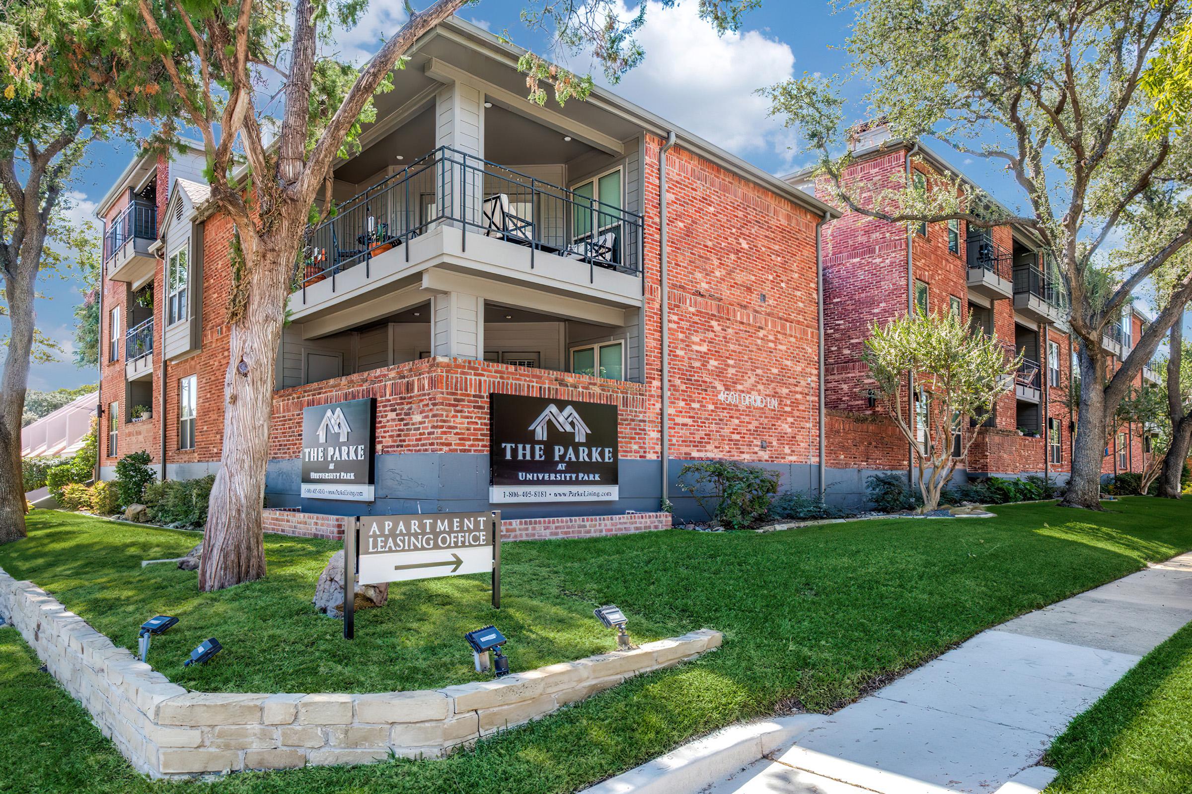 A modern brick apartment building with balconies, featuring a leasing office sign pointing left. Surrounding green lawns and trees enhance the inviting atmosphere of the residential complex. The sunny sky adds to the appealing environment of the area.