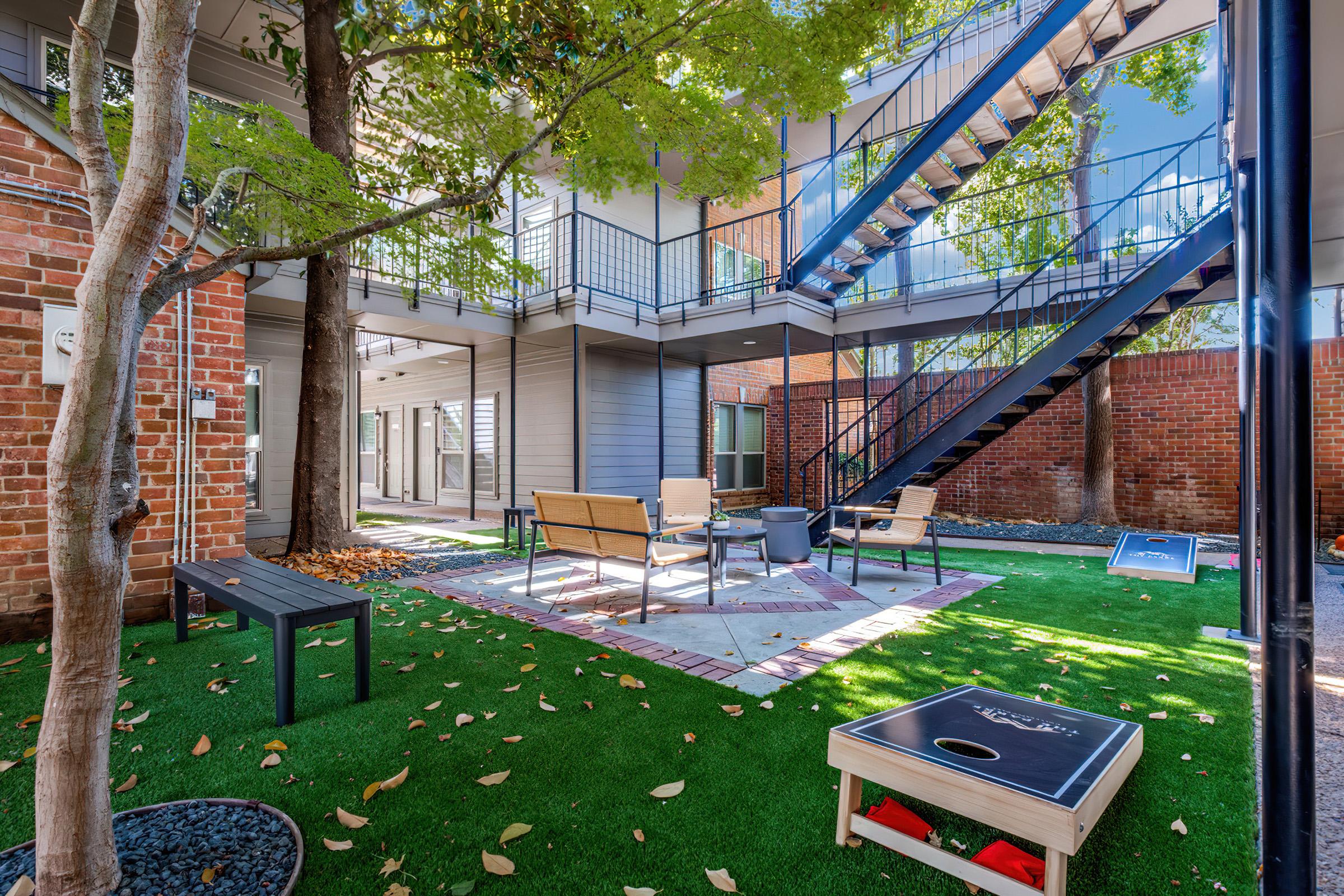 A well-landscaped courtyard features a cozy seating area with chairs and a table, surrounded by artificial grass and trees. A blue fire escape staircase leads to upper levels, while a cornhole game is set up nearby. The space is bright and inviting, providing a relaxed outdoor gathering place.